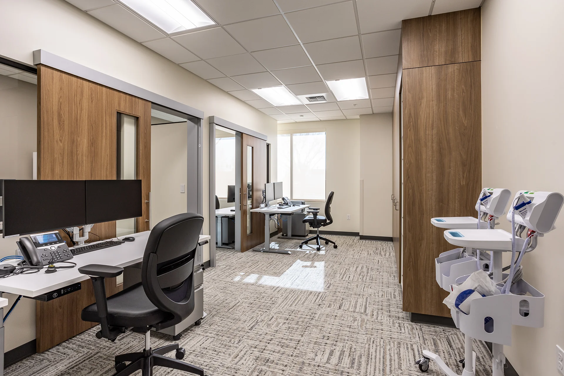 Professional office suite for medical professionals showcasing modular white desks, dual-monitor desktop configurations, ergonomic black mesh chairs, custom floor-to-ceiling wood grain storage wardrobes, low-pile patterned carpet tiles, and mobile medical carts.