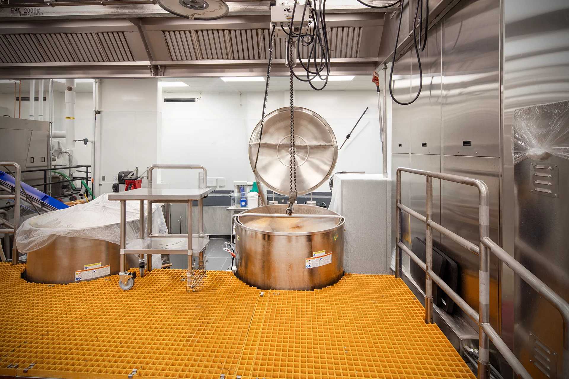 Commercial kitchen installation highlighting a large capacity stainless steel mixing kettle with an open lid suspended by a mechanical hoist system, set against stainless steel wall cladding and durable yellow safety floor grating.