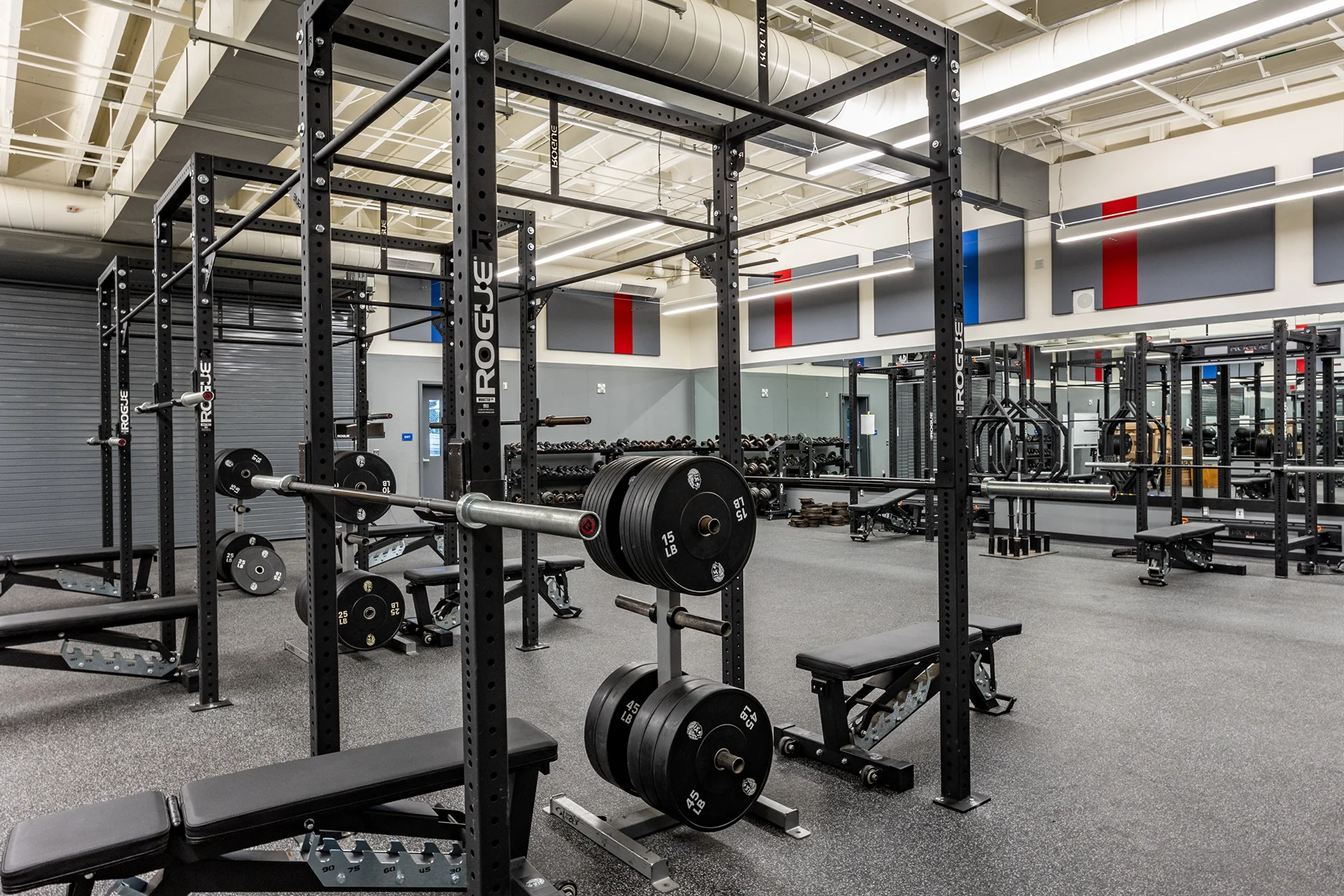 Modern commercial-grade high school weight room highlighting black steel Rogue power lifting racks, custom weight plate storage trees with rubber plates, thick black rubber athletic flooring, and wall-mounted perimeter mirrors.