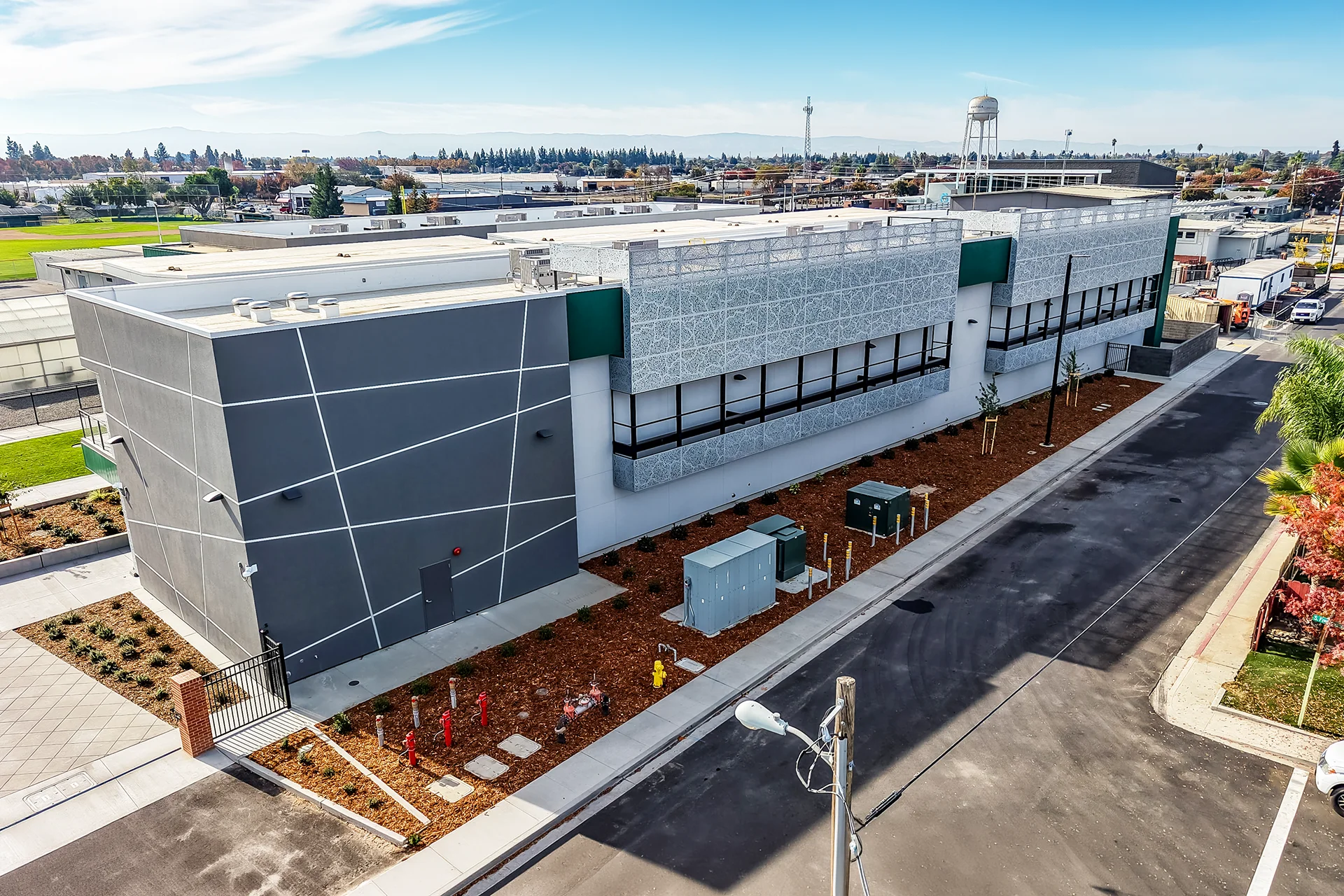 Overhead drone perspective of a modern multi-material institutional building facade in Manteca, California, looking down at a paved asphalt fire lane, pad-mounted utility infrastructure boxes, a decorative block safety wall, and distant valley mountain horizons under a clear blue sky.
