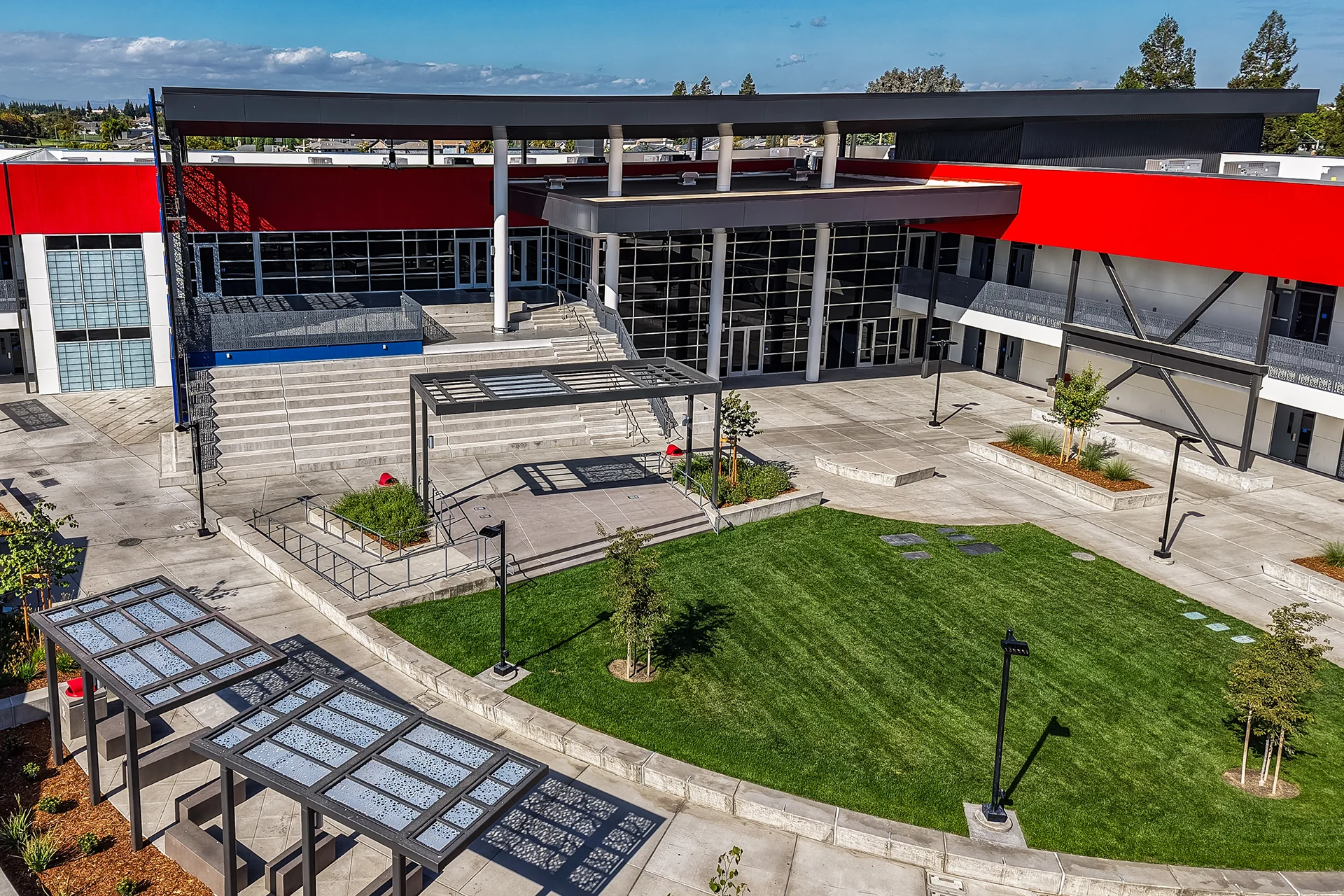 Elevated drone view of a newly constructed institutional campus courtyard highlighting a manicured grass lawn, multi-tiered concrete plaza steps, steel shade pergolas, modern black light poles, and a glass storefront building facade.