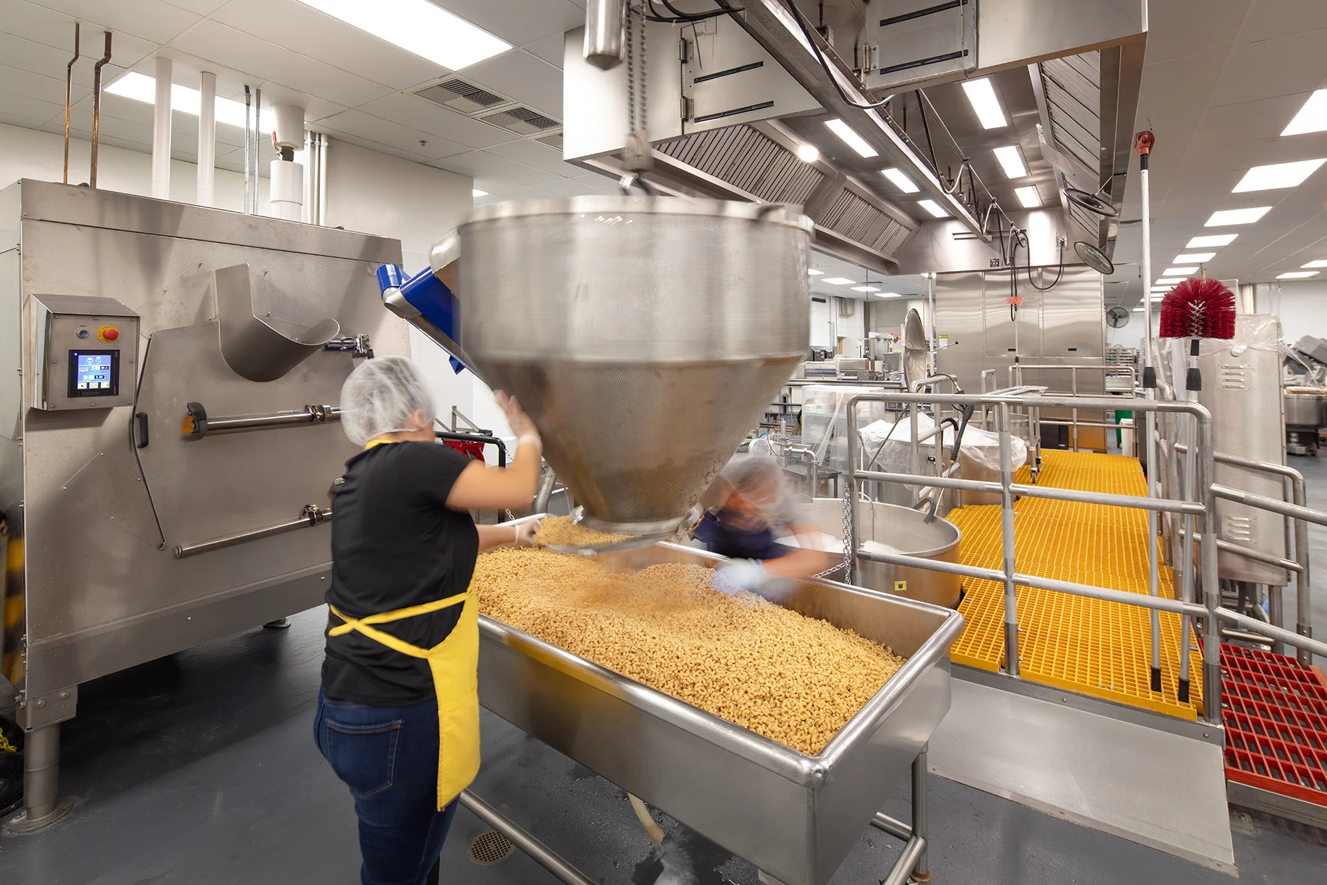 Food service production line installation showcasing an overhead stainless steel hopper discharging bulk ingredients into a large stainless steel trough, featuring slip resistant yellow grating floors and stainless steel wash down machinery.