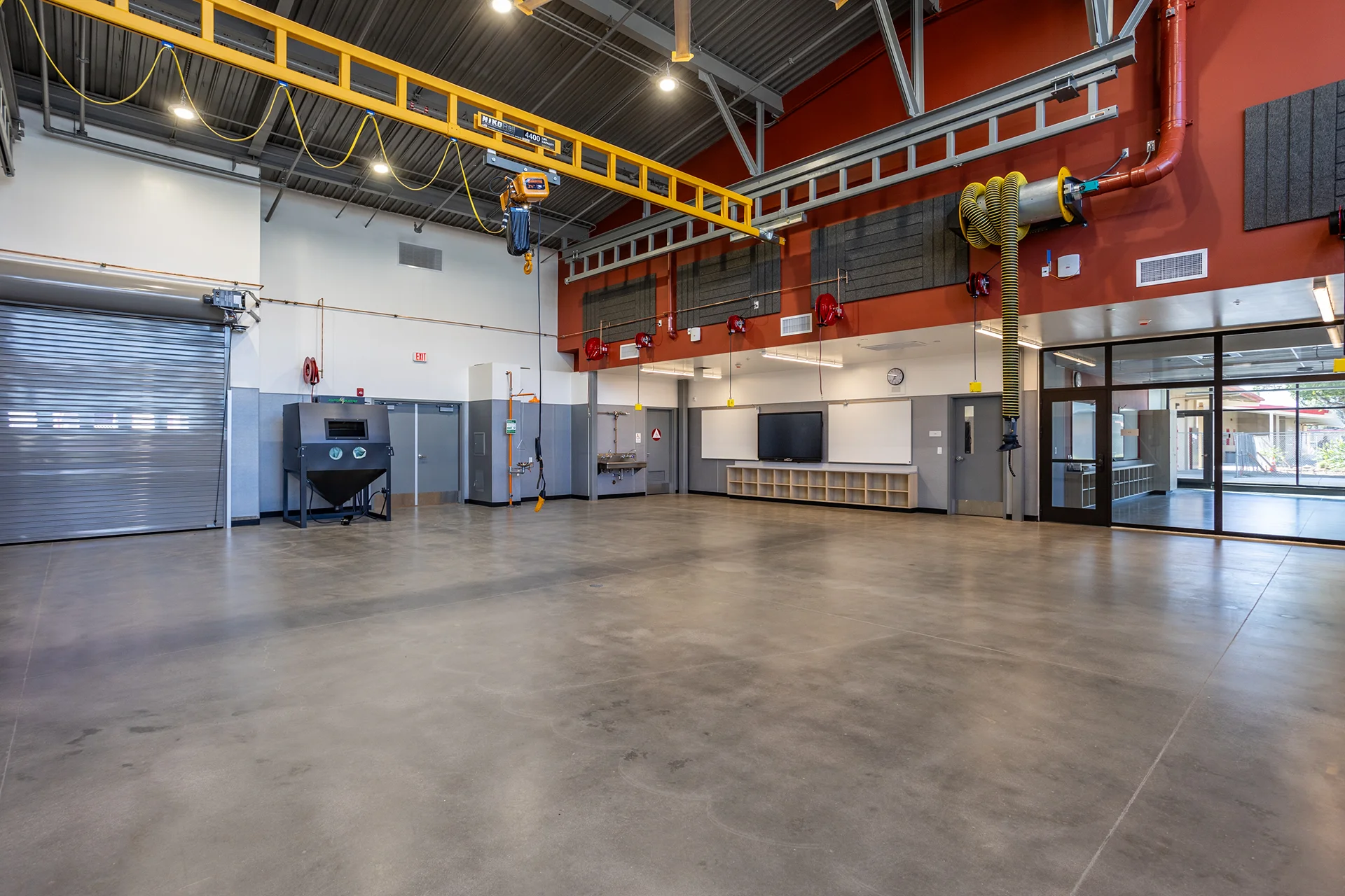 Large-scale overview of an expansive vocational training bay showing polished concrete floors, a yellow overhead crane rail system, wall-mounted flexible source-capture exhaust arms, acoustic sound baffling, and a glass-walled observation office.
