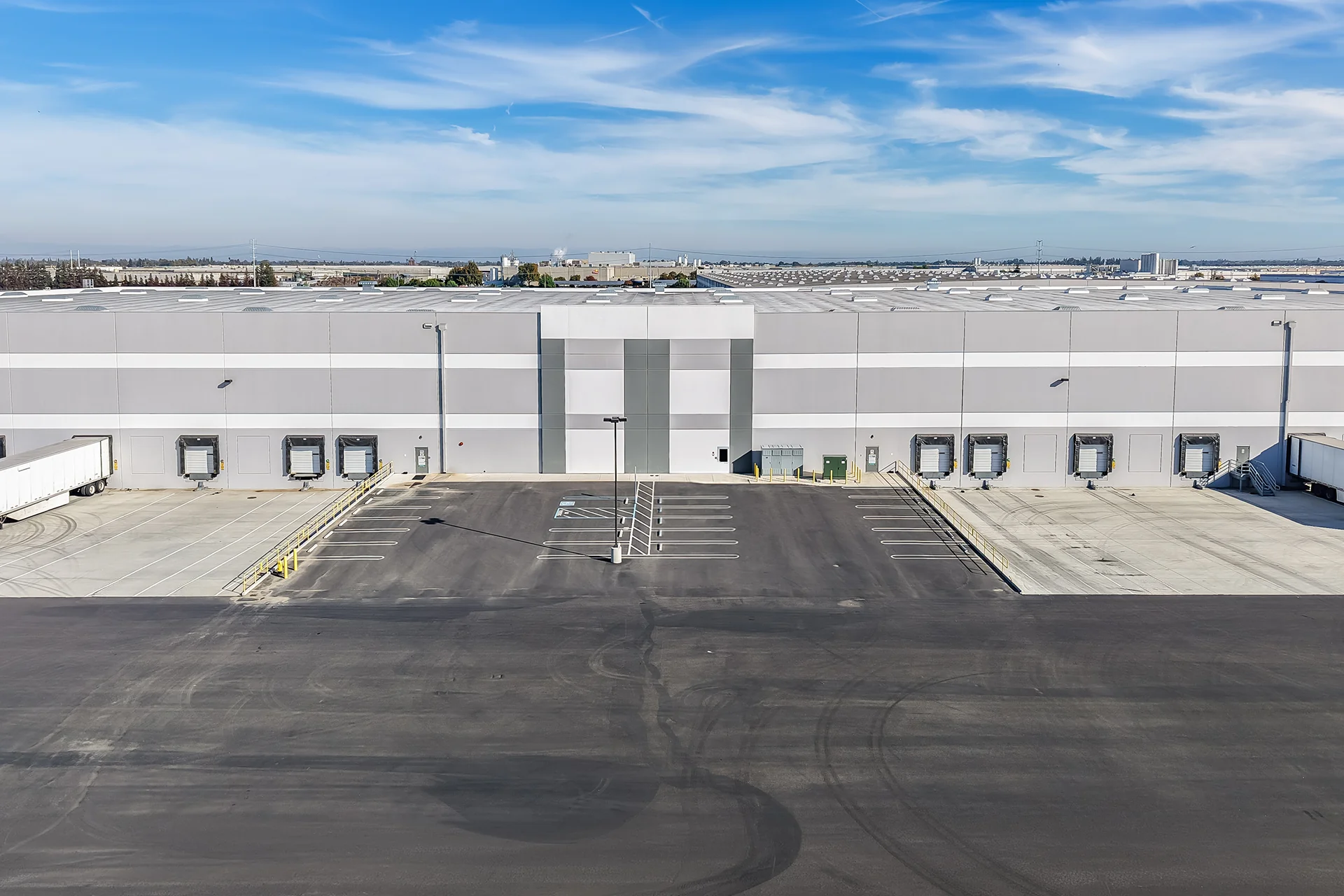 Close-up aerial view of a cross-dock logistics building facade showing insulated sectional overhead doors, protective rubber dock seals, a cast-in-place concrete terminal apron with safety pipe railings, and center-aligned tractor-trailer parking stalls.