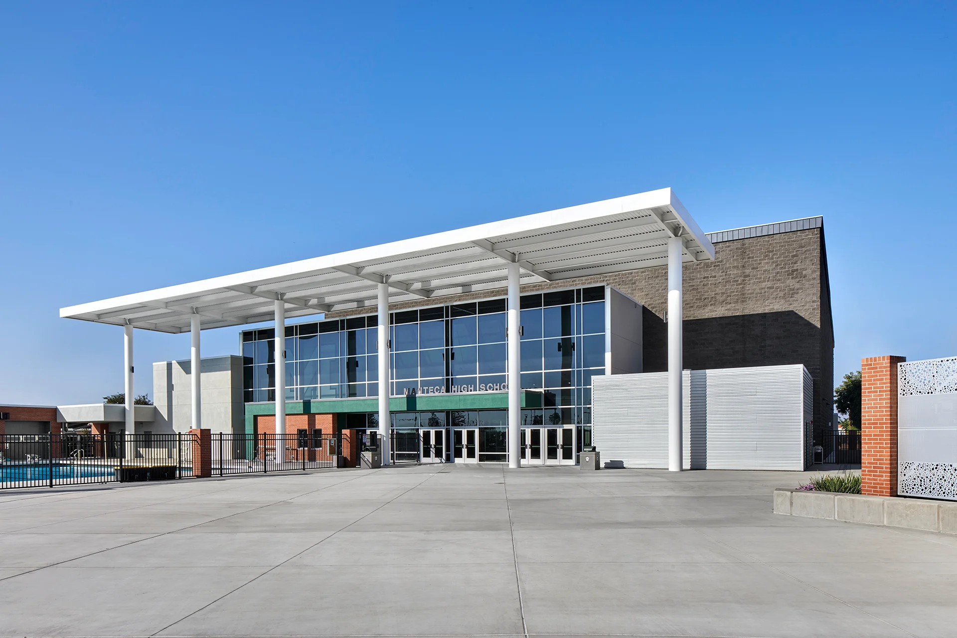 Wide architectural shot focusing on a multi-story educational building entryway plaza, highlighting clean-lined clear aluminum window walls, integrated green fascia bands, structural masonry brick piers, black industrial iron gates, and extensive concrete hardscaping.