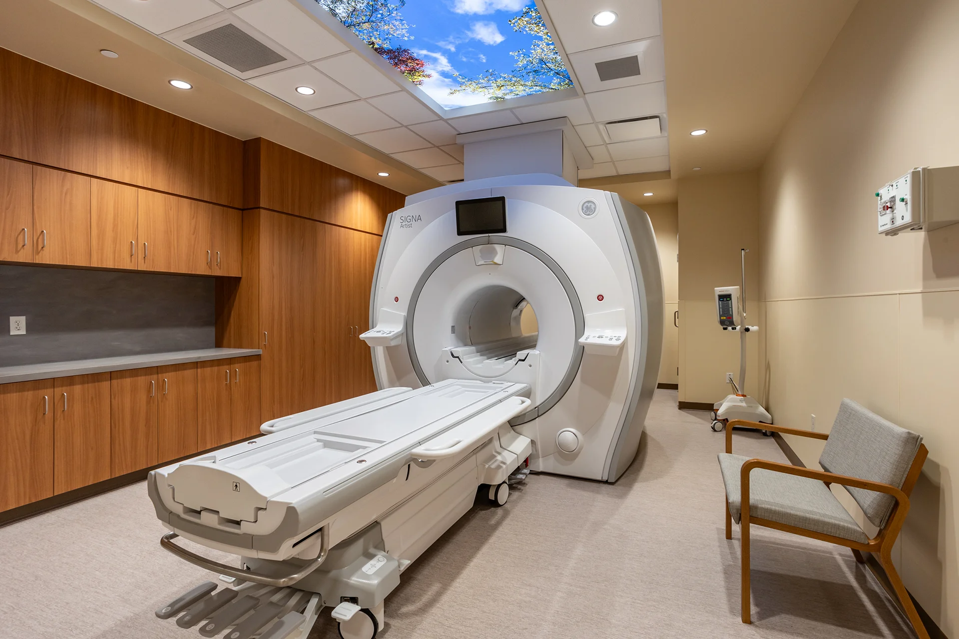 Wide-angle view of a medical diagnostic room featuring a GE SIGNA Artist MRI system with an extended patient bed, a mobile contrast injector stand, built-in wooden cabinetry, an armchair, and integrated acoustic wall paneling.