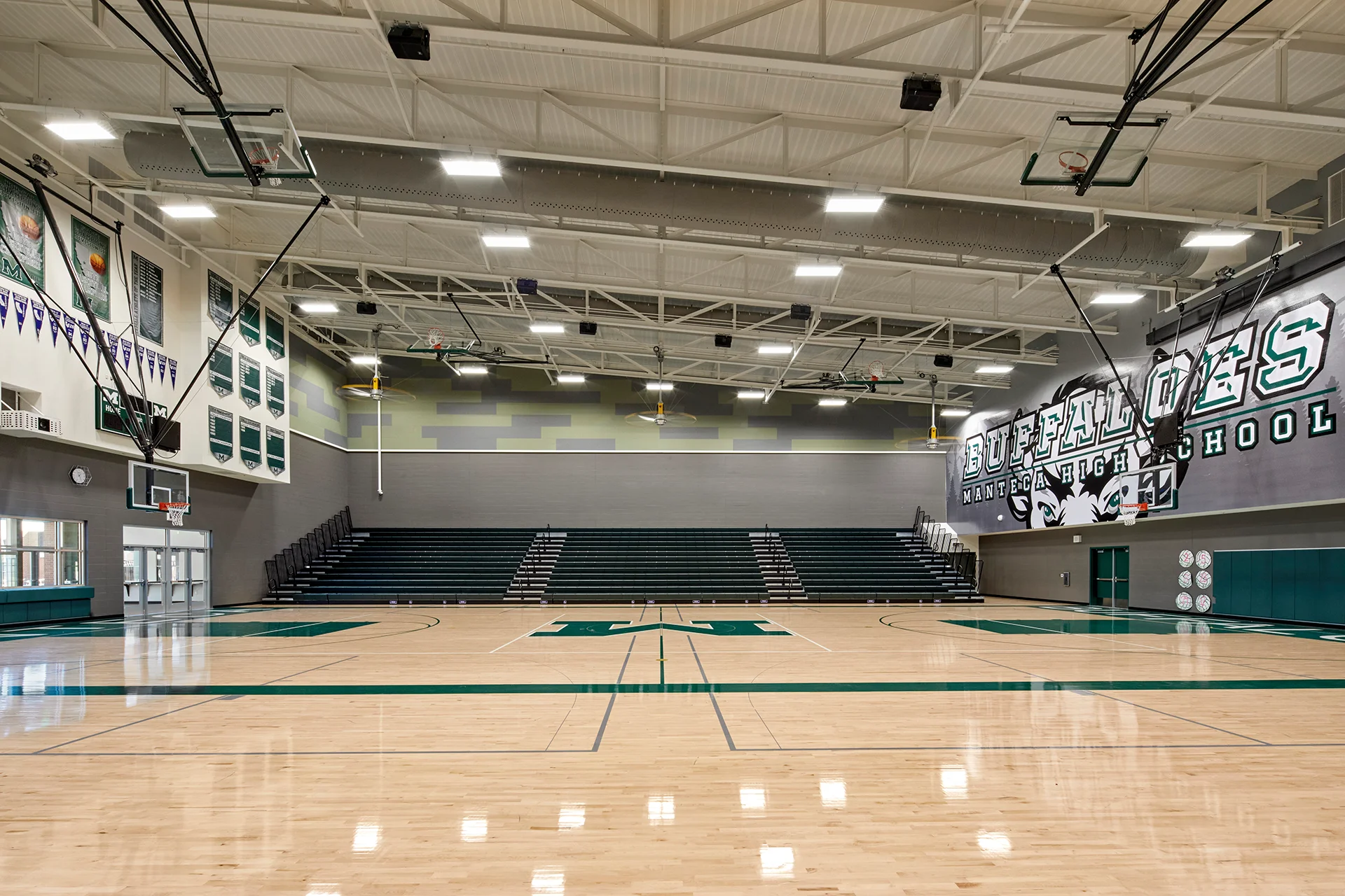 Full interior perspective of a newly constructed institutional basketball gymnasium, displaying a high-gloss polished maple court with green boundary lines, tiered green spectator bleachers, retractable glass backboards, structural steel roof trusses, and a large custom graphic mural.