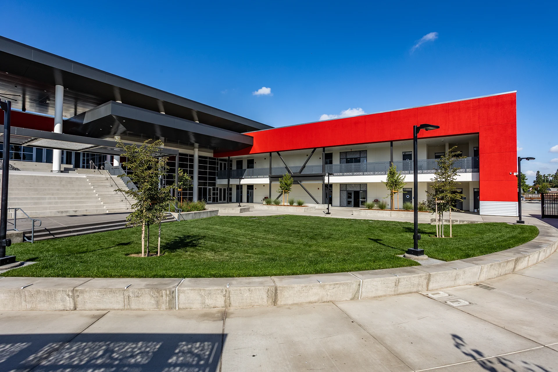 Modern high school campus courtyard featuring a manicured green lawn bordered by a low concrete retaining wall. The adjacent multi-story classroom building shows a bold red stucco facade accent, an upper-level pedestrian walkway with grey perforated metal railings, steel cross-bracing, and black LED pole lighting fixtures under a clear blue sky.