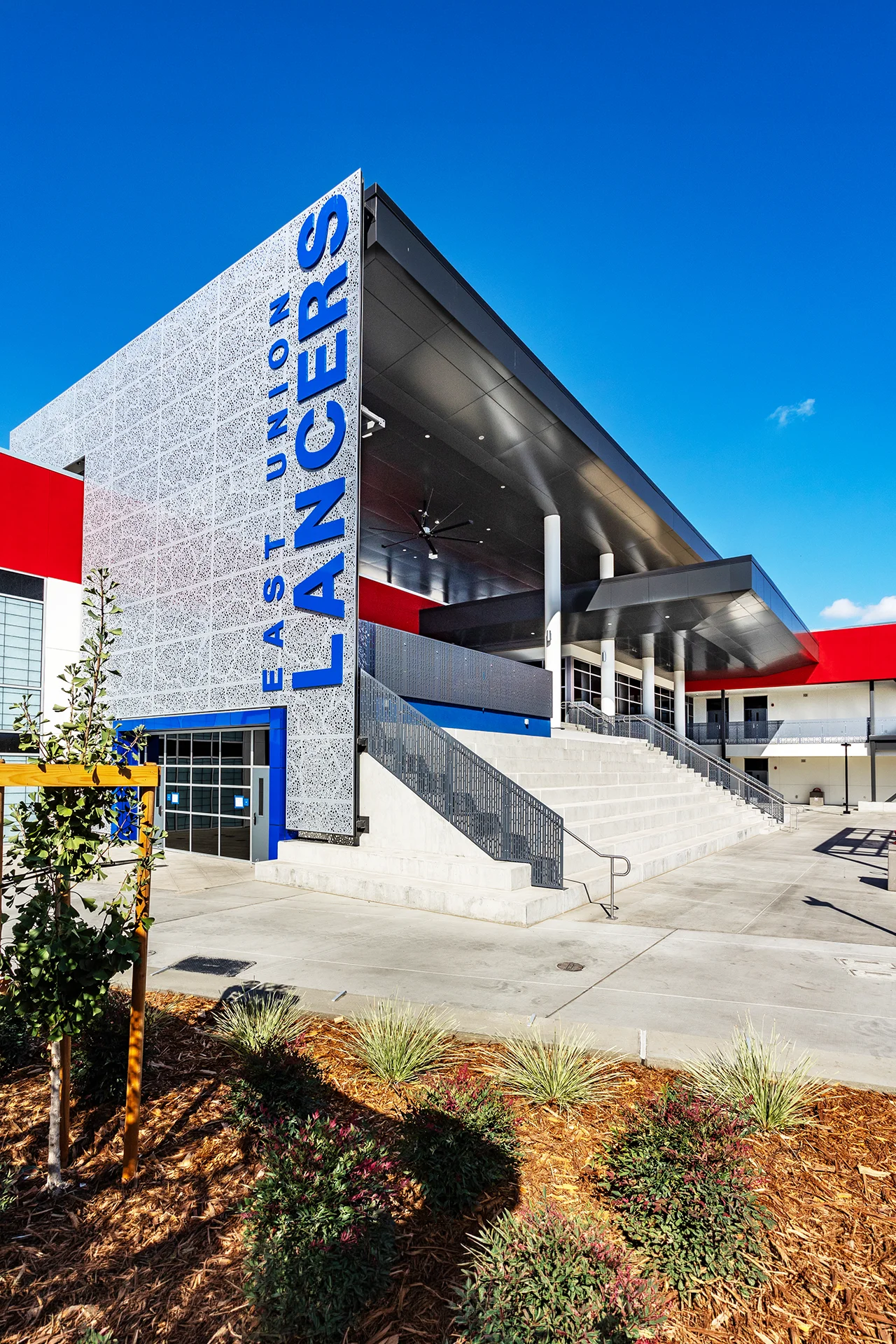 Dynamic architectural shot showcasing wide concrete amphitheater steps leading up to an elevated campus deck area under a dark metal canopy soffit. The structural design features a large patterned white metal screen feature wall with vertical blue East Union Lancers lettering and a grey metal protective safety railing.