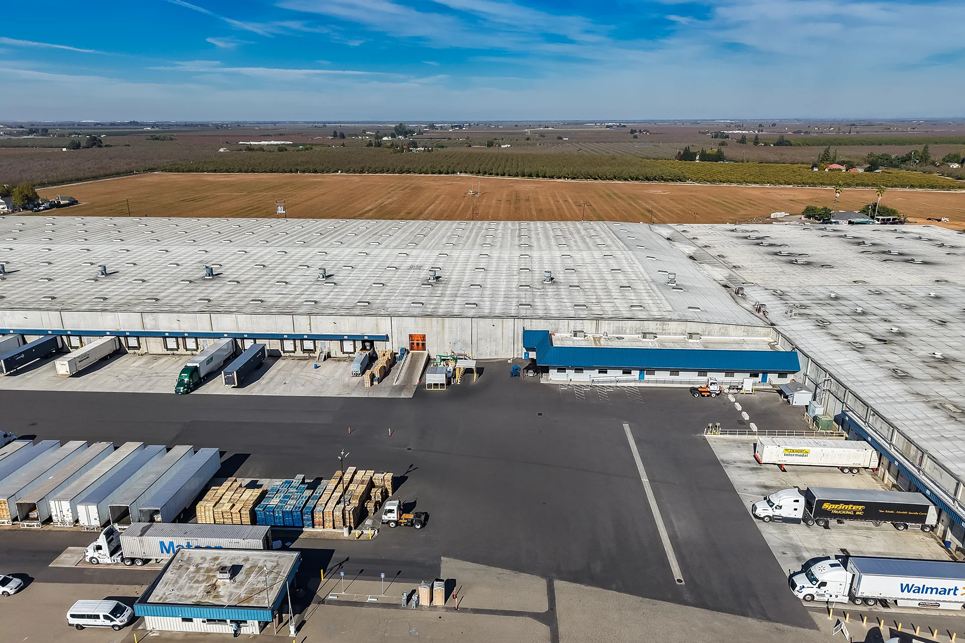 Elevated drone shot of a food-grade industrial warehouse complex, highlighting a cast-in-place concrete terminal apron, a prominent blue-roofed administrative annex, a striped employee parking zone, multiple consumer fleet trailers including Walmart and Sprinter models, and regional transport infrastructure.