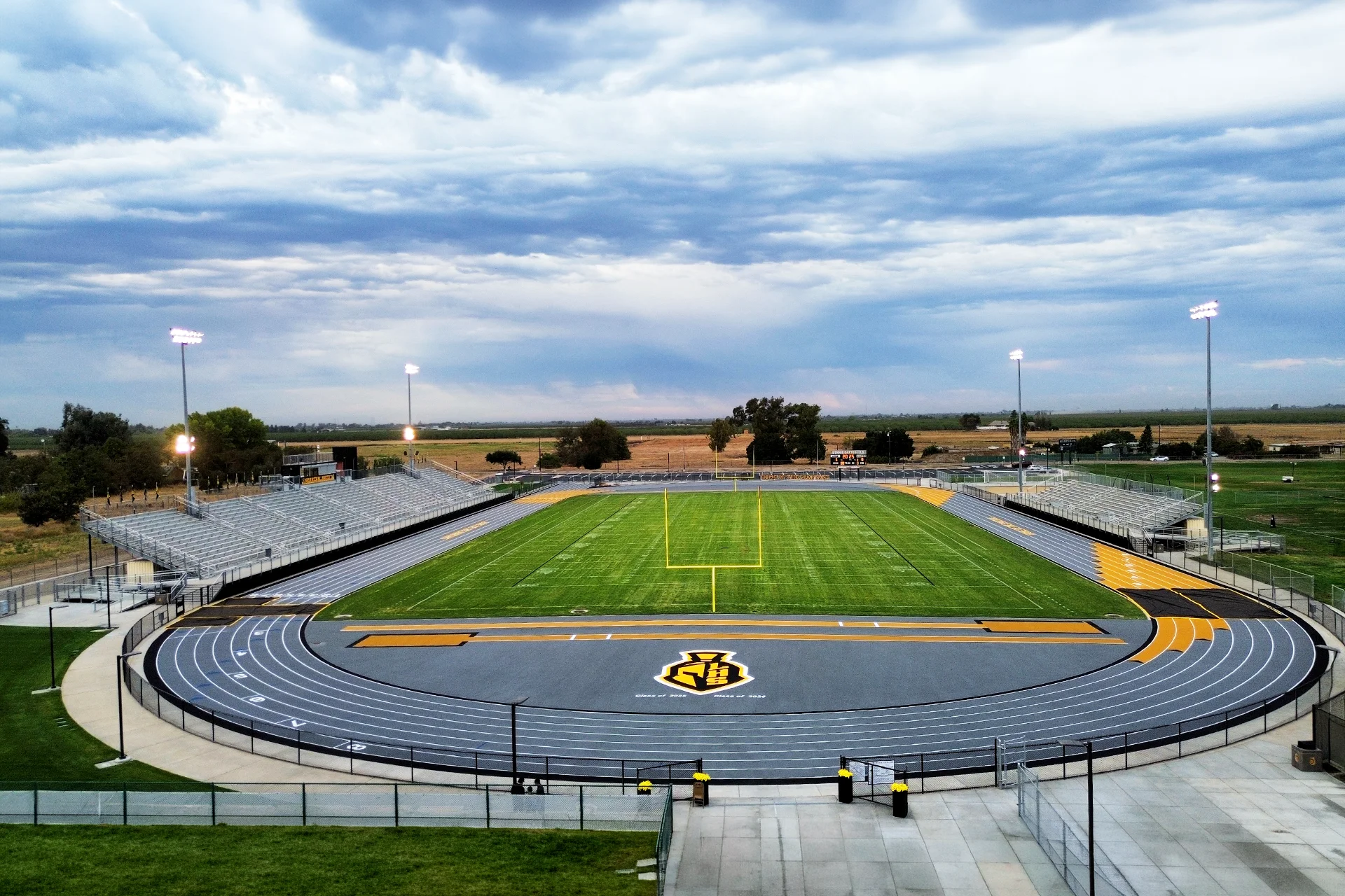 An elevated stadium overview of the green football turf, yellow goalposts, and a grey rubberized track under active stadium light poles.
