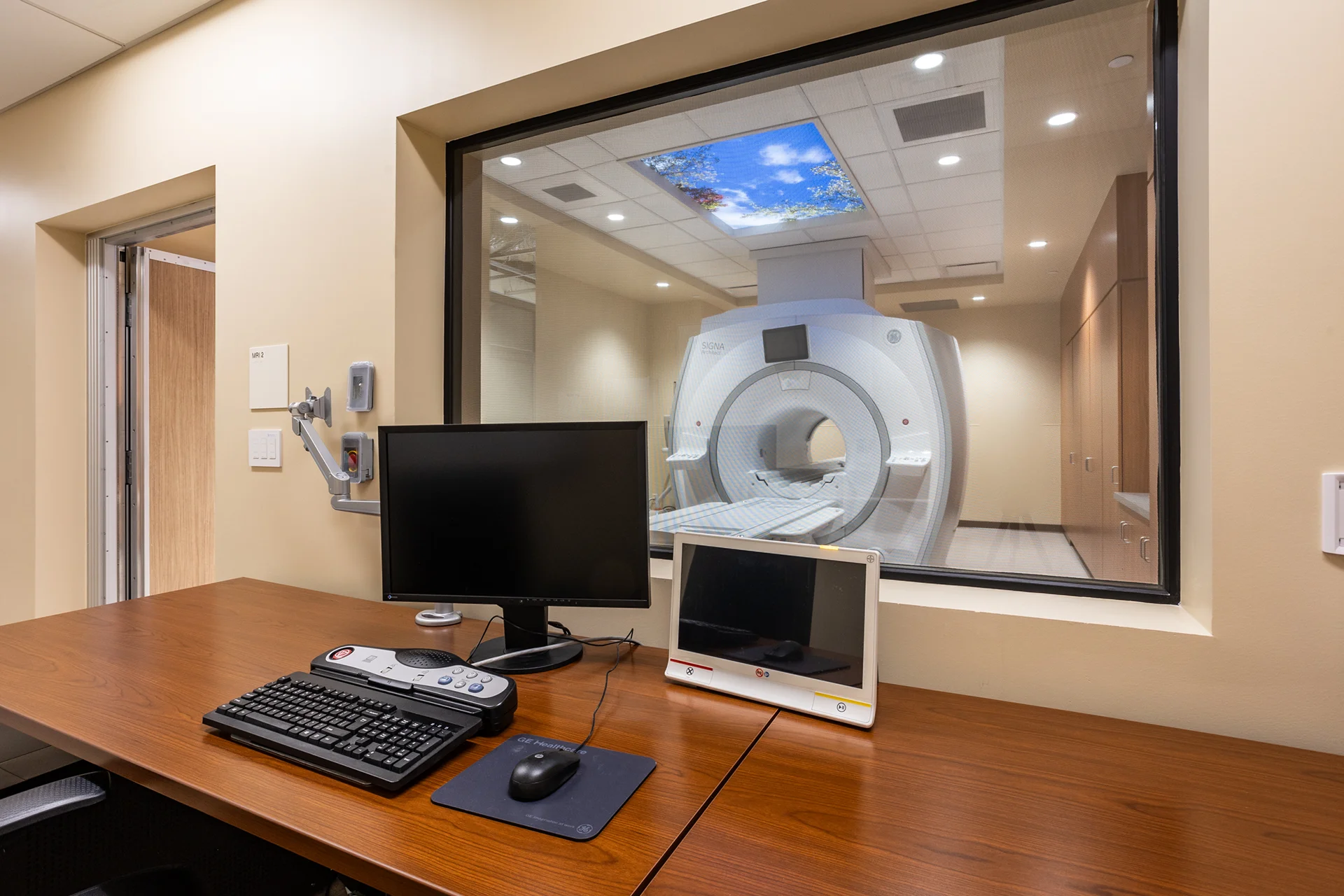 Operator workstation inside a medical diagnostics lab featuring computer peripherals, a desktop intercom module, and an expansive observation window providing clear lines of sight to a central scanner unit.