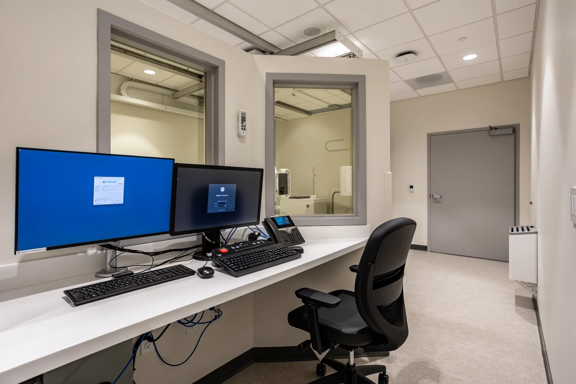 Dedicated radiology technician control station featuring a wide white work counter with multiple imaging monitors, specialized control panels, and double-paned lead-glass viewing windows looking directly into an adjacent diagnostic X-ray room.