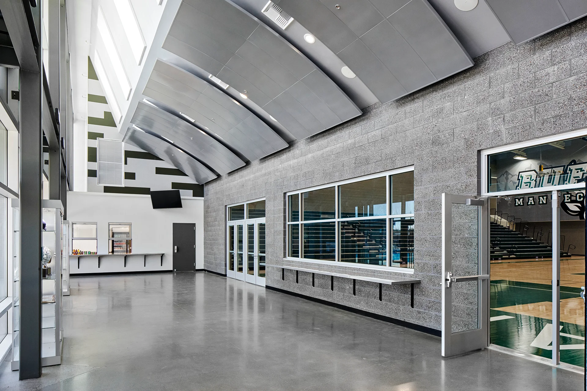 Interior view of a high school gymnasium lobby in San Joaquin County, featuring reflective polished concrete flooring, a split-face concrete masonry unit wall with commercial transaction windows, an integrated concession counter stand, and custom curved metal sound-dampening ceiling baffles.