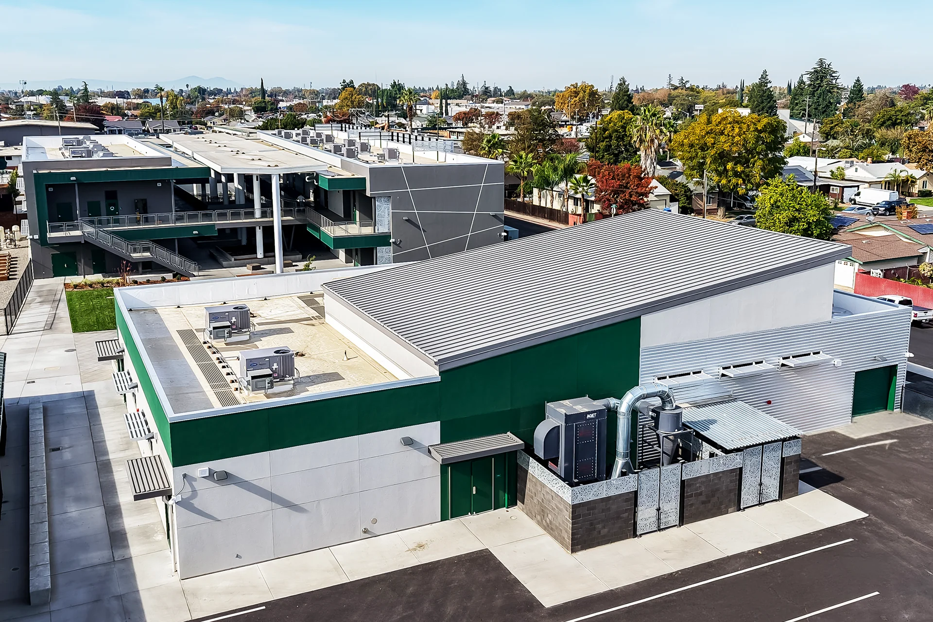 Elevated bird's-eye view looking down on a newly constructed school building with a standing seam metal roof section, a flat TPO roof area housing commercial Carrier package unit air conditioners, and a ground-level industrial dust collection system enclosed by a CMU wall.
