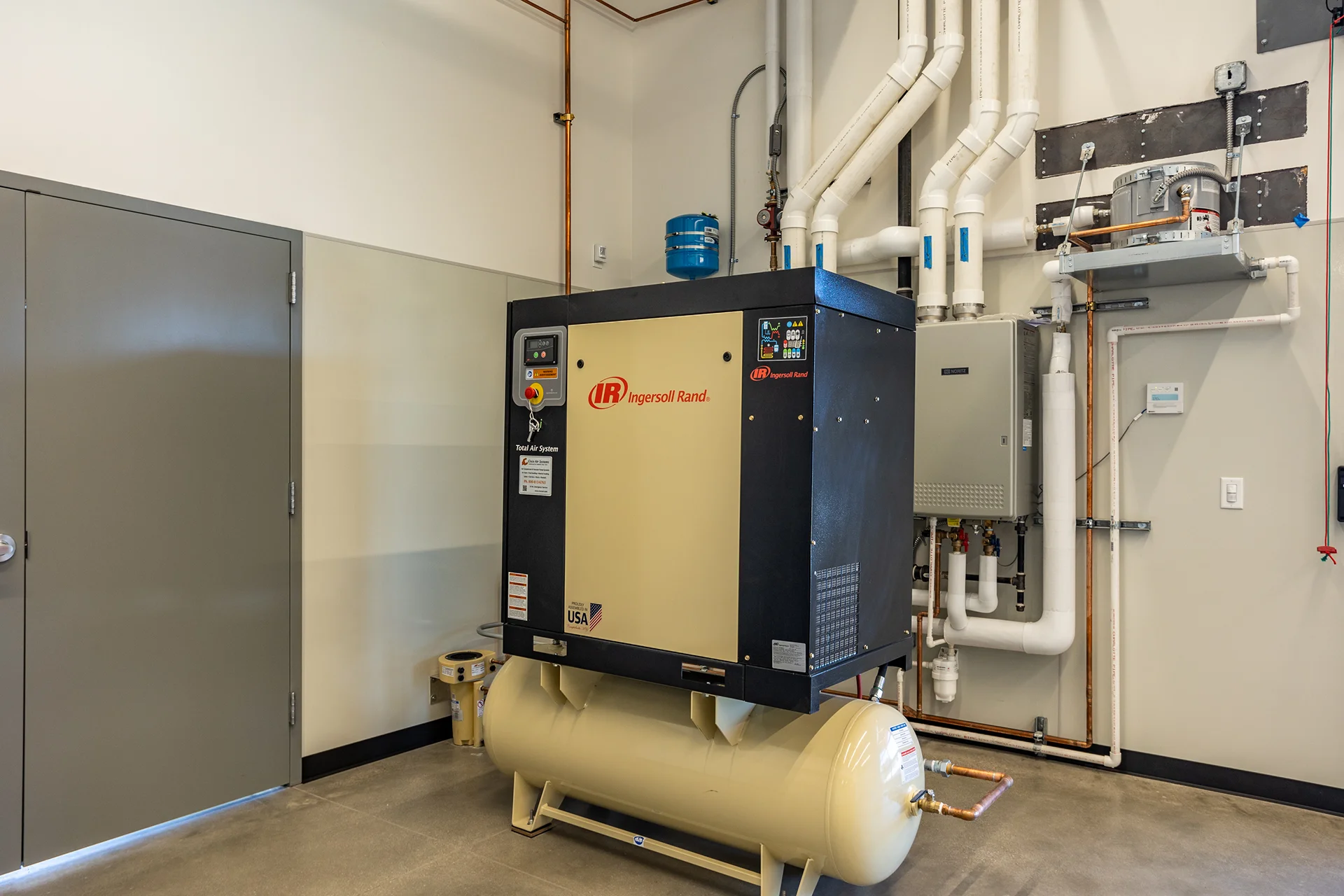 Mechanical equipment corner featuring a large enclosed air compressor unit on a cream-colored receiver tank, flanked by commercial wall-mounted boilers, high-voltage electrical runs, and structured PVC drain lines over finished concrete.