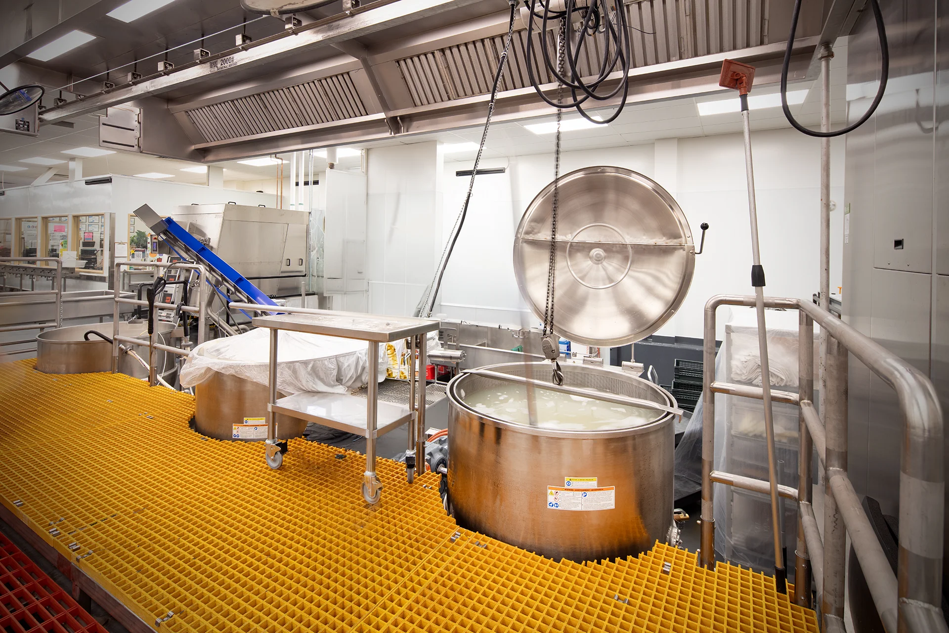 Mechanical view of an institutional kitchen cooking line highlighting a large capacity boiling kettle, overhead utility support structures, hoist cables, and custom structural steel platforms with yellow safety walkway grating.