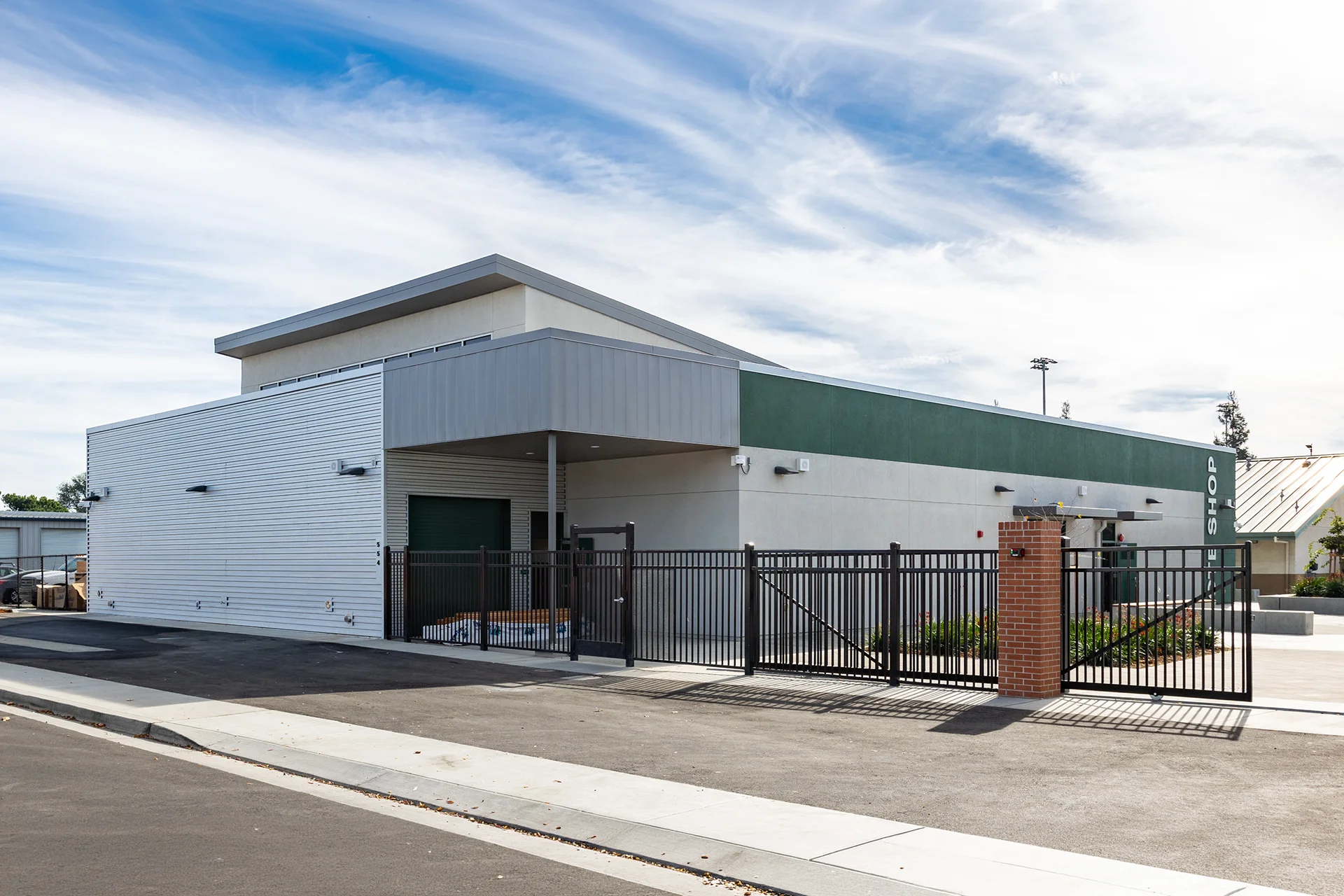Exterior view of a school vocational or maintenance facility in Manteca, California, displaying clean white horizontal corrugated metal siding, a flat roofline with metal coping, black steel security fencing, an open gate, and an integrated masonry brick pillar entry.