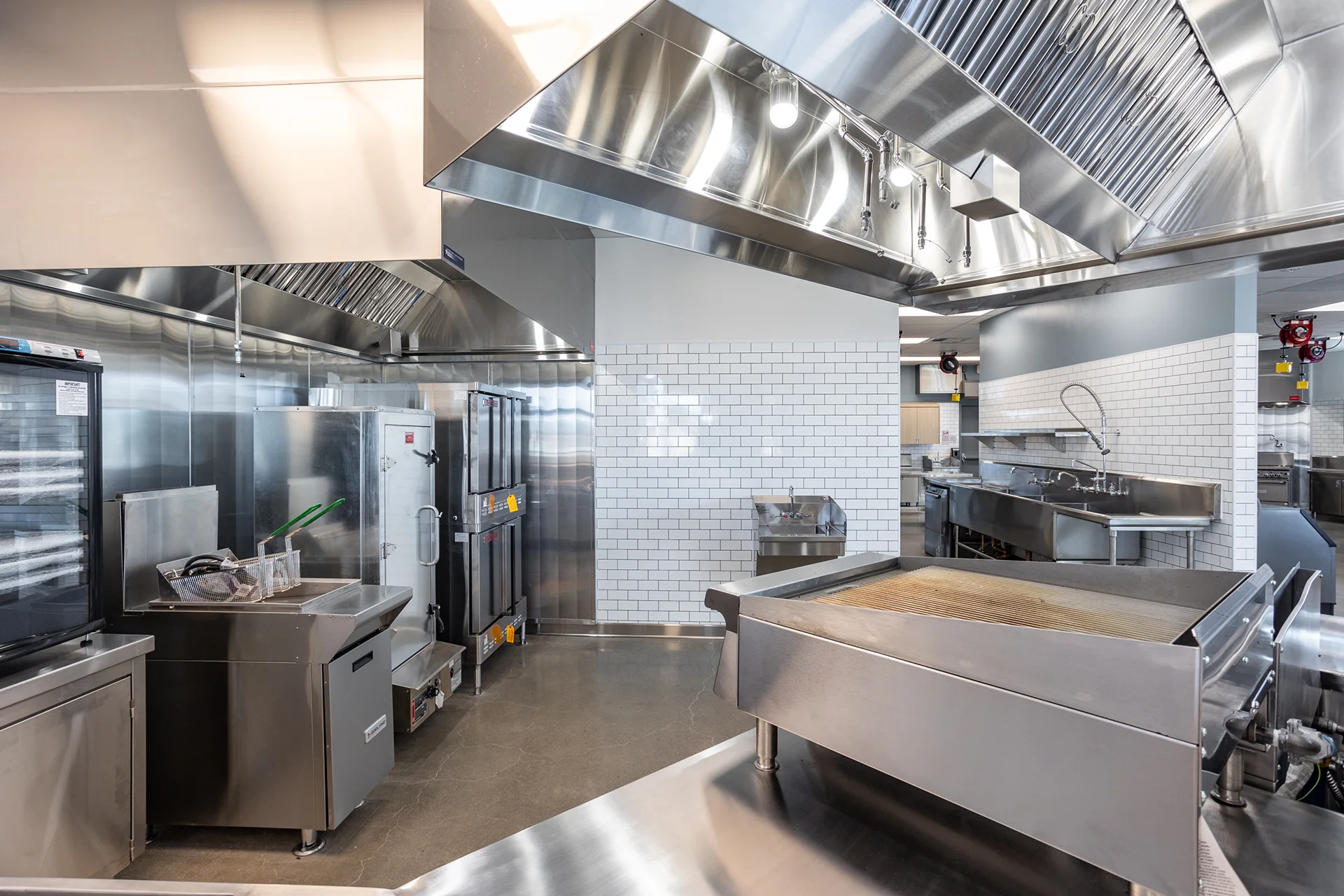 Interior profile view of a high-volume food service line featuring stainless steel commercial ovens, a gas deep fryer, a large open-top griddle station, and custom floor-to-ceiling polished stainless wall cladding.