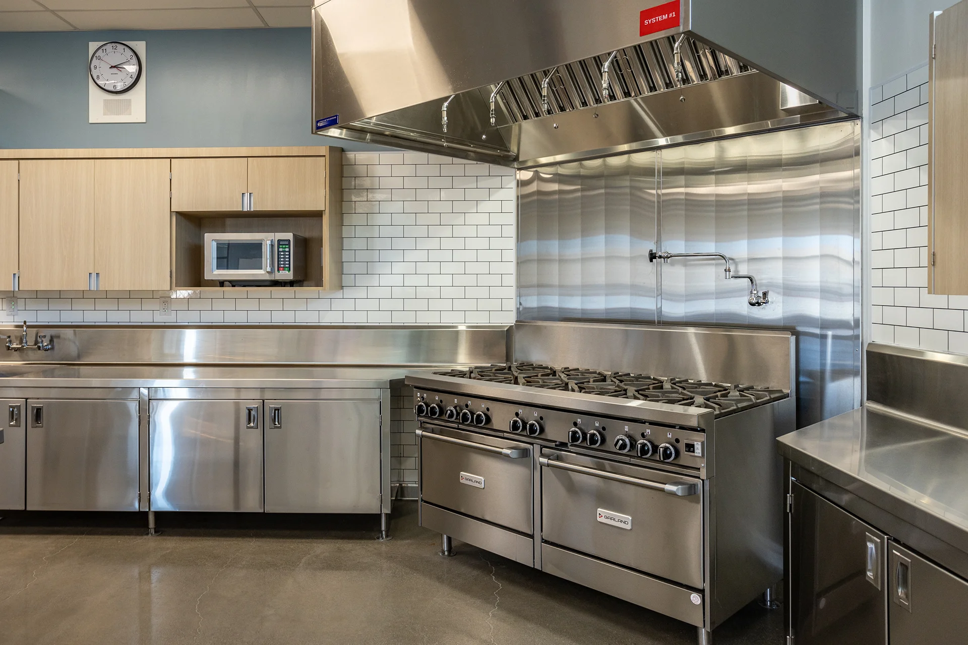 Medium shot of a professional cooking line highlighting a commercial gas range, matching stainless steel under-counter storage compartments, a wall-mounted analog clock, and clean white subway tile surfacing.
