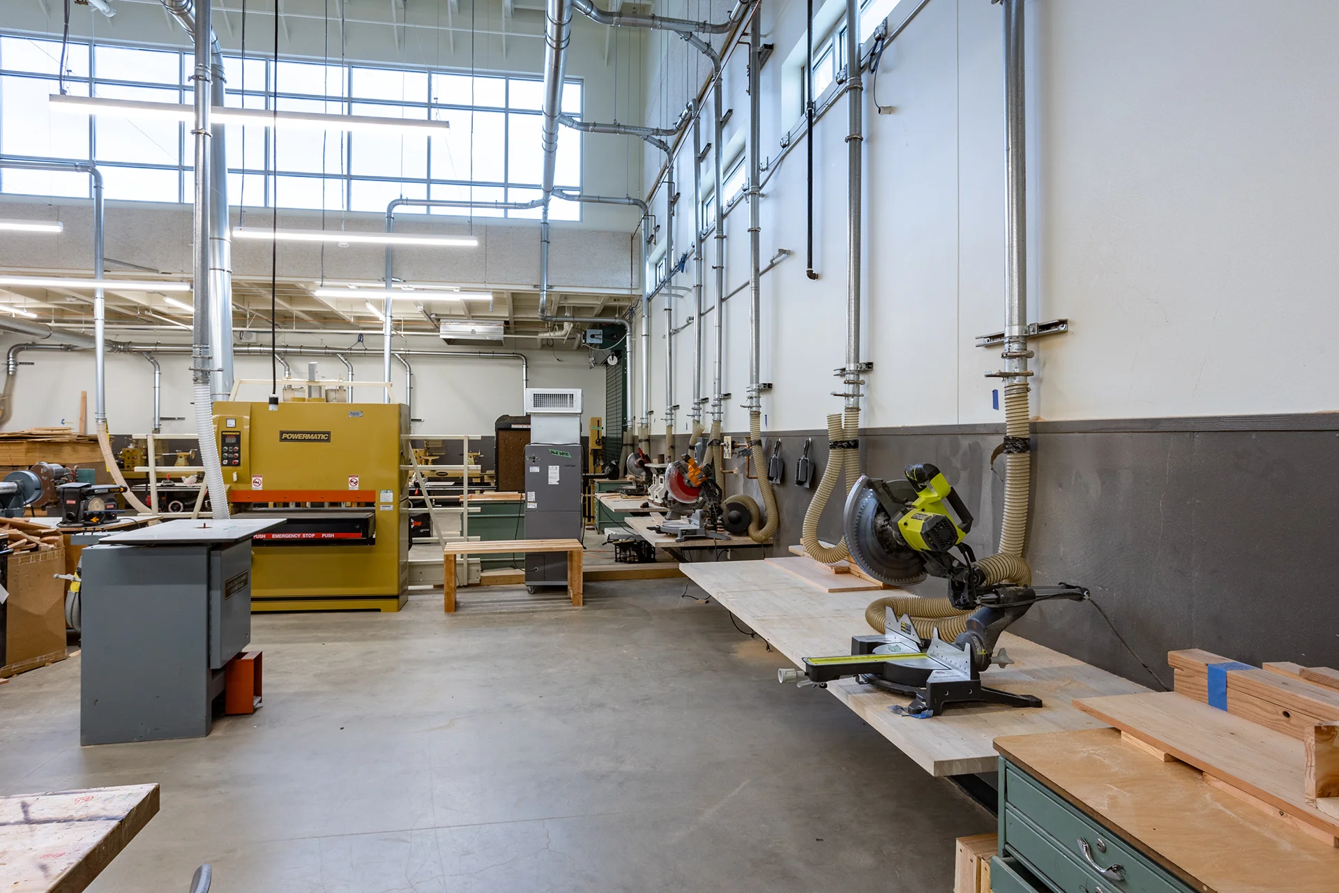 Detailed interior view of a school carpentry workshop, highlighting a sequence of Ryobi and miter saws mounted on long plywood counter extensions along a grey-wainscoted wall, featuring dedicated flexible vacuum hoses connected to a main source-capture ventilation system.