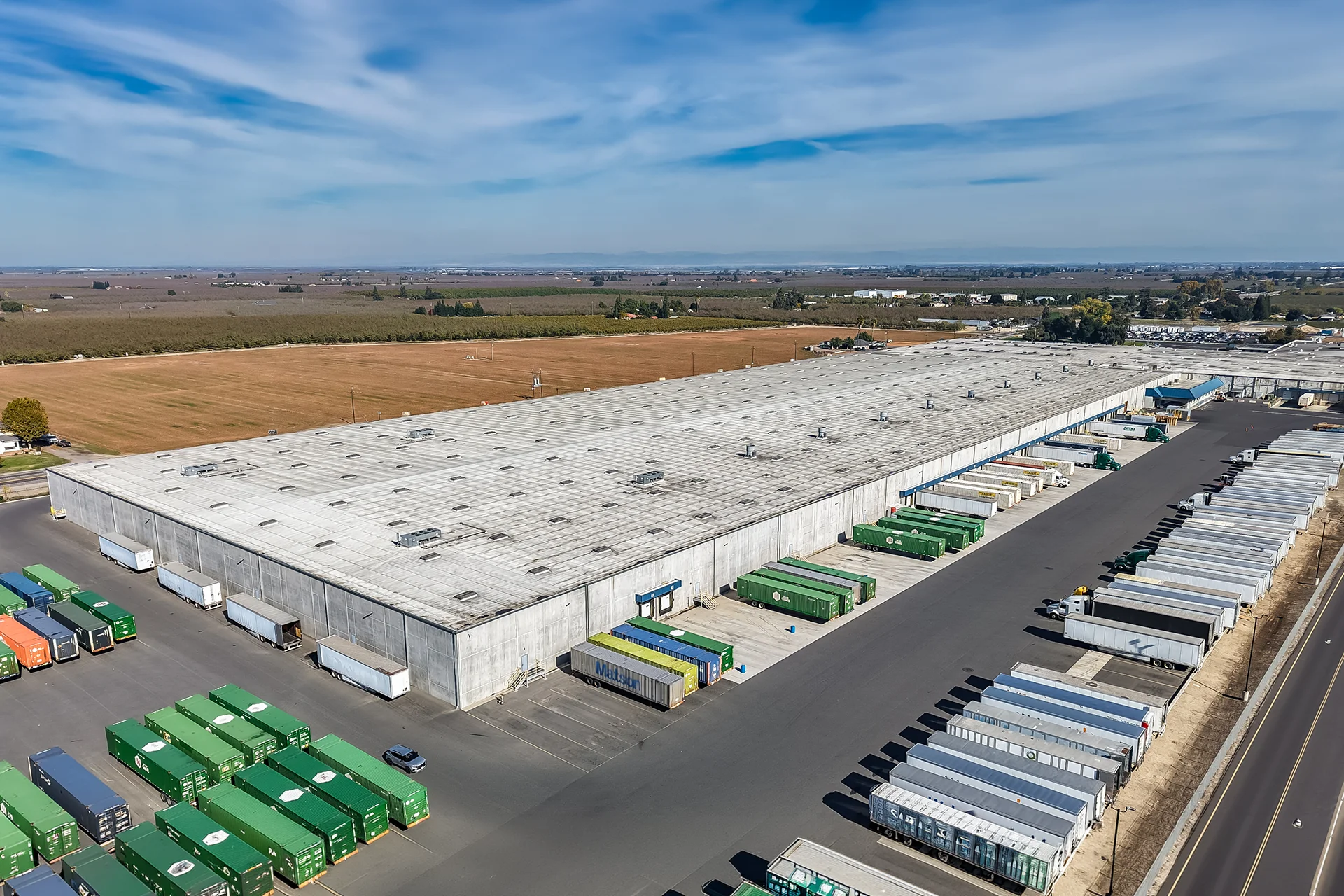 Three-quarter aerial perspective of an institutional tilt-up concrete logistics building, showing deep concrete trailer dolly pads, numerous green and blue intermodal shipping containers, a fenced security perimeter, and clear sightlines across Central Valley agricultural land.