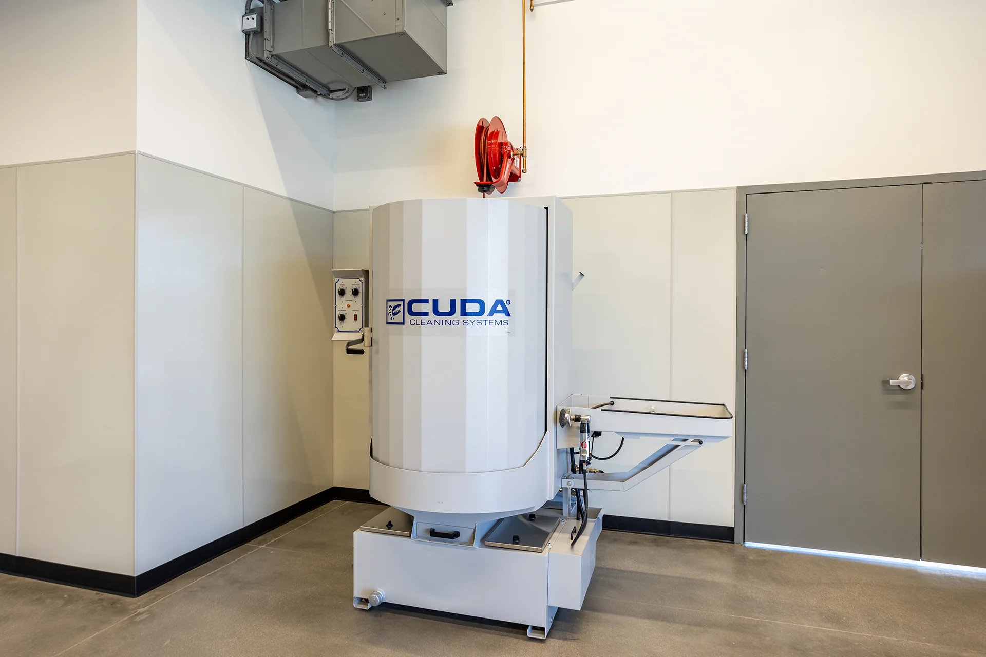 Heavy-duty automated parts degreasing system installed on a smooth concrete floor in a workshop corner, showcasing rigid wall protection panels, a ceiling-mounted red hose reel, and code-compliant gray steel utility doors.