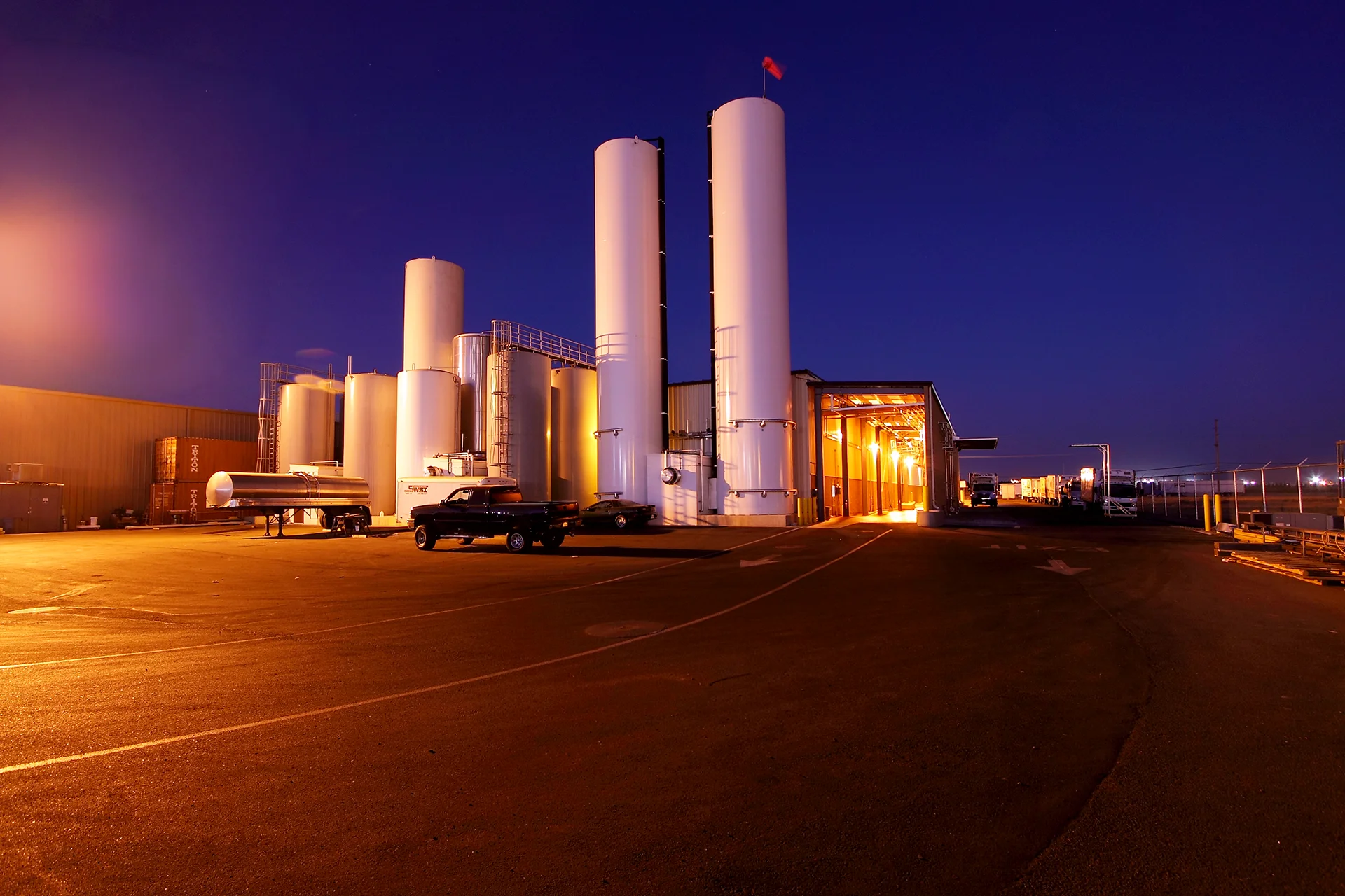 Nighttime perspective of a food manufacturing plant highlighting a large cluster of vertical storage tanks illuminated by exterior safety lighting, an open-sided covered equipment staging area with bright overhead fixtures, and transport trailers parked on the surrounding asphalt compound.