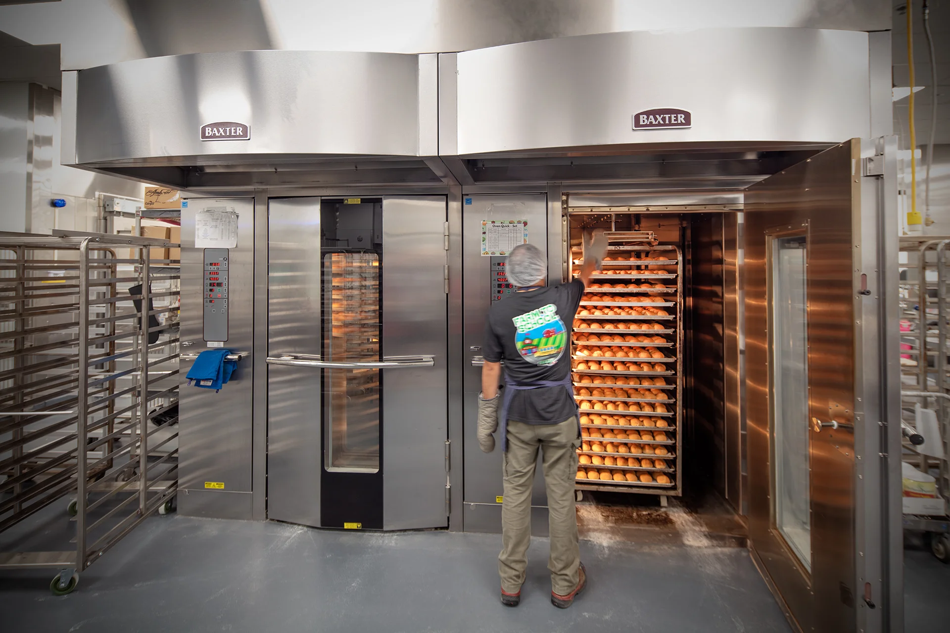A kitchen worker loading a mobile stainless steel tray rack filled with baked goods into a commercial Baxter rotating rack oven inside a large scale institutional food production facility in Sacramento County.