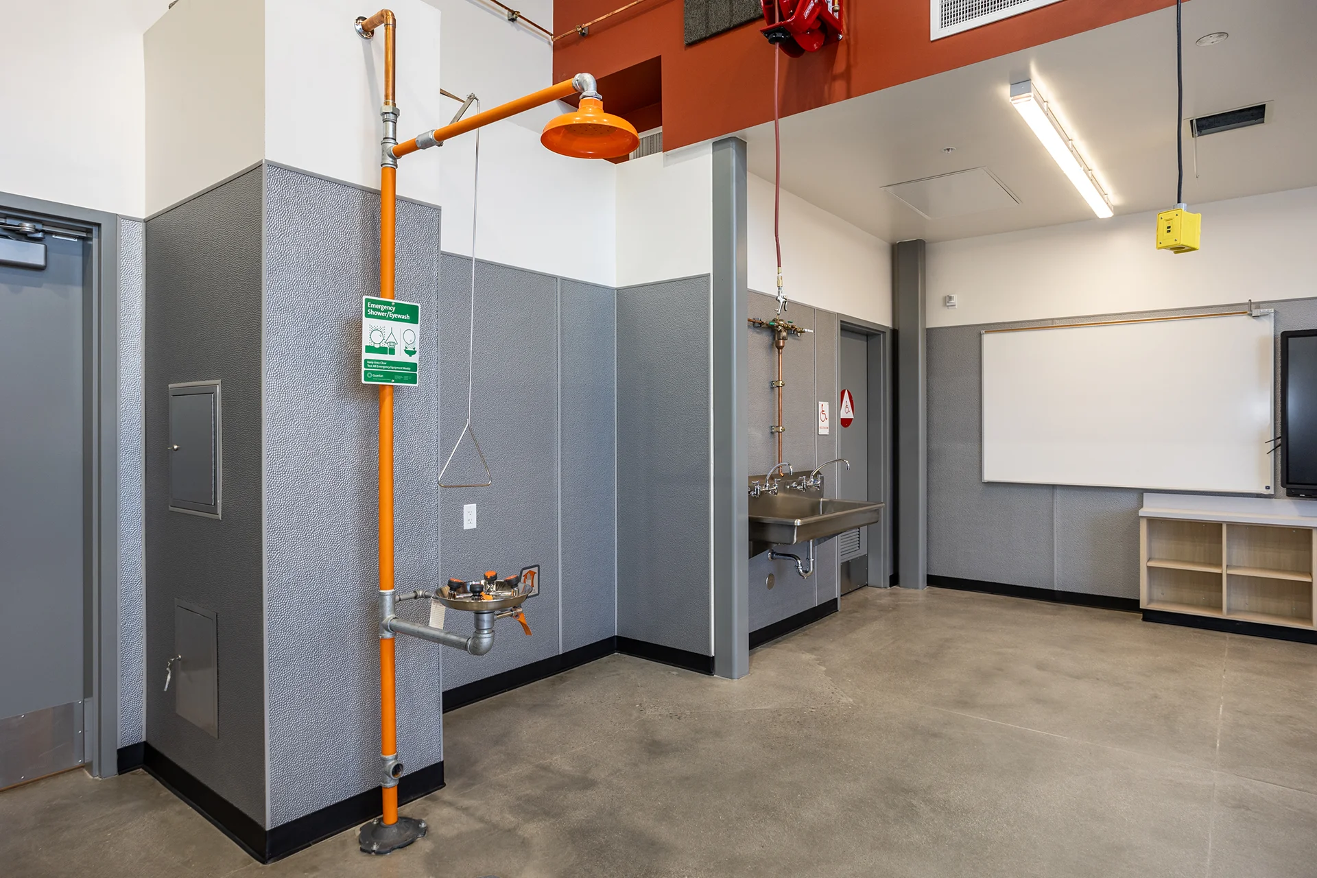 High-visibility orange emergency eyewash and overhead deluge shower fixture installed with rigid copper piping against textured gray moisture-resistant wall panels, positioned adjacent to an ADA-compliant stainless steel handwashing trough.