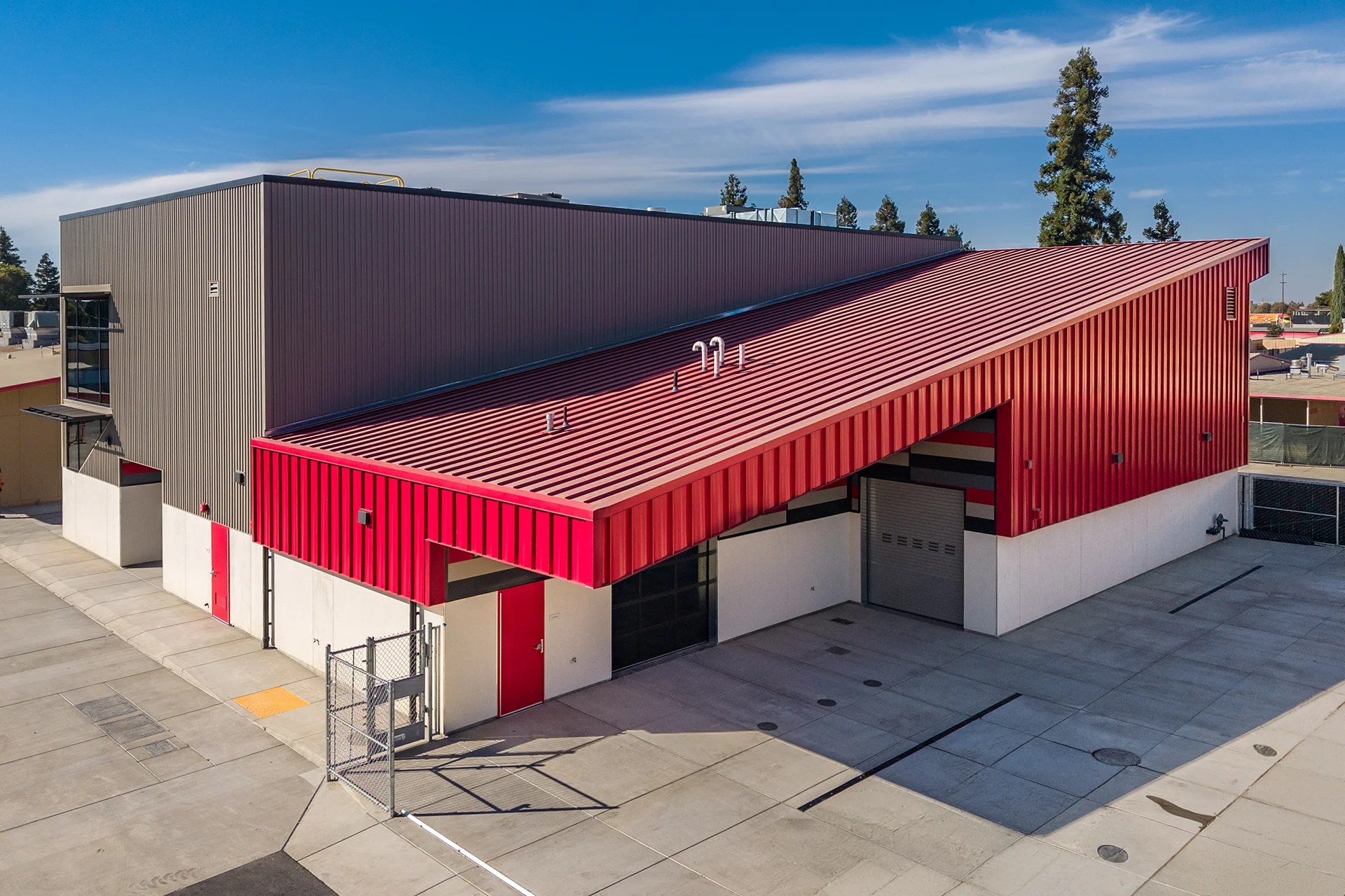 Elevated exterior view of a technical facility showcasing a red metal roof awning extending over an outdoor loading apron, integrated commercial roll-up garage doors, crimson personnel exit doors, and chain-link security fencing under a clear sky.