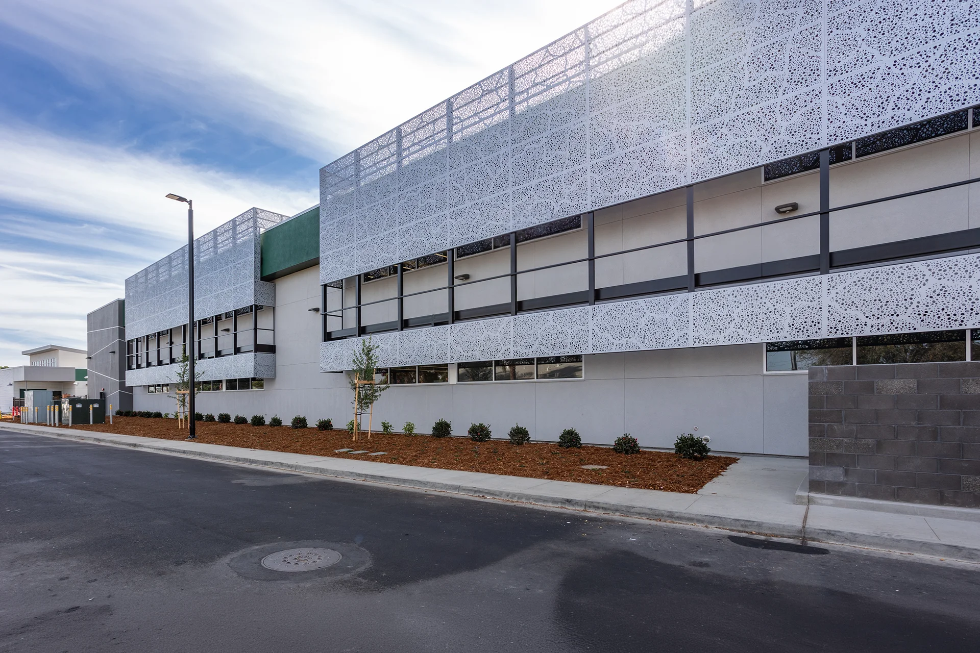 Wide shot looking down a paved campus roadway beside a contemporary educational building in Manteca, California, highlighting extensive perforated metal facade screening, integrated exterior LED lighting, freshly laid bark mulch landscaping with small shrubs, and a decorative concrete block wall.
