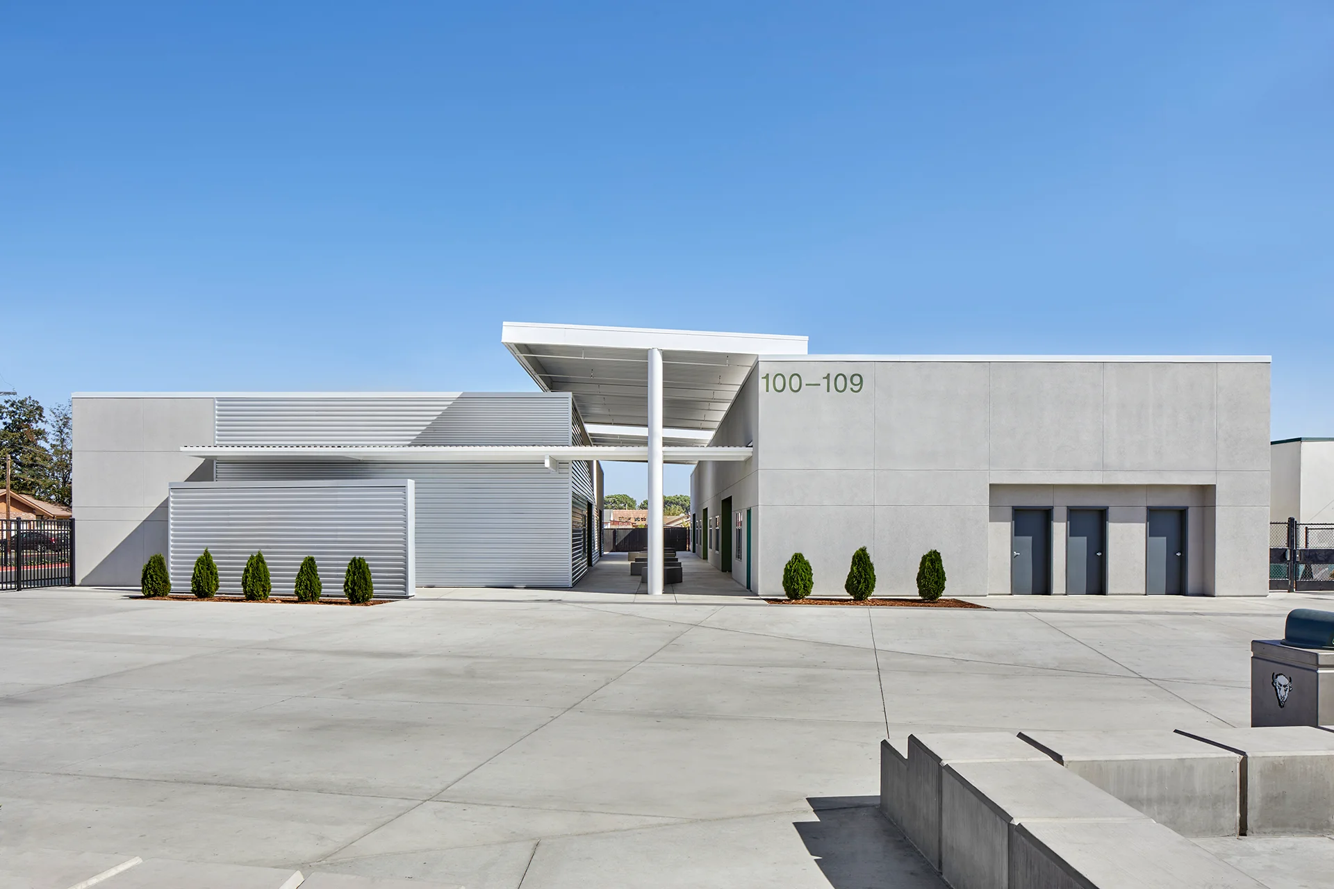 Clear daytime view of a modern school facility entryway in Manteca, California, showcasing smooth grey stucco structures balanced by silver horizontal corrugated metal paneling, geometric concrete walkways, a high-clearance canopy roof, and neat rows of manicured conical boxwood shrubs.