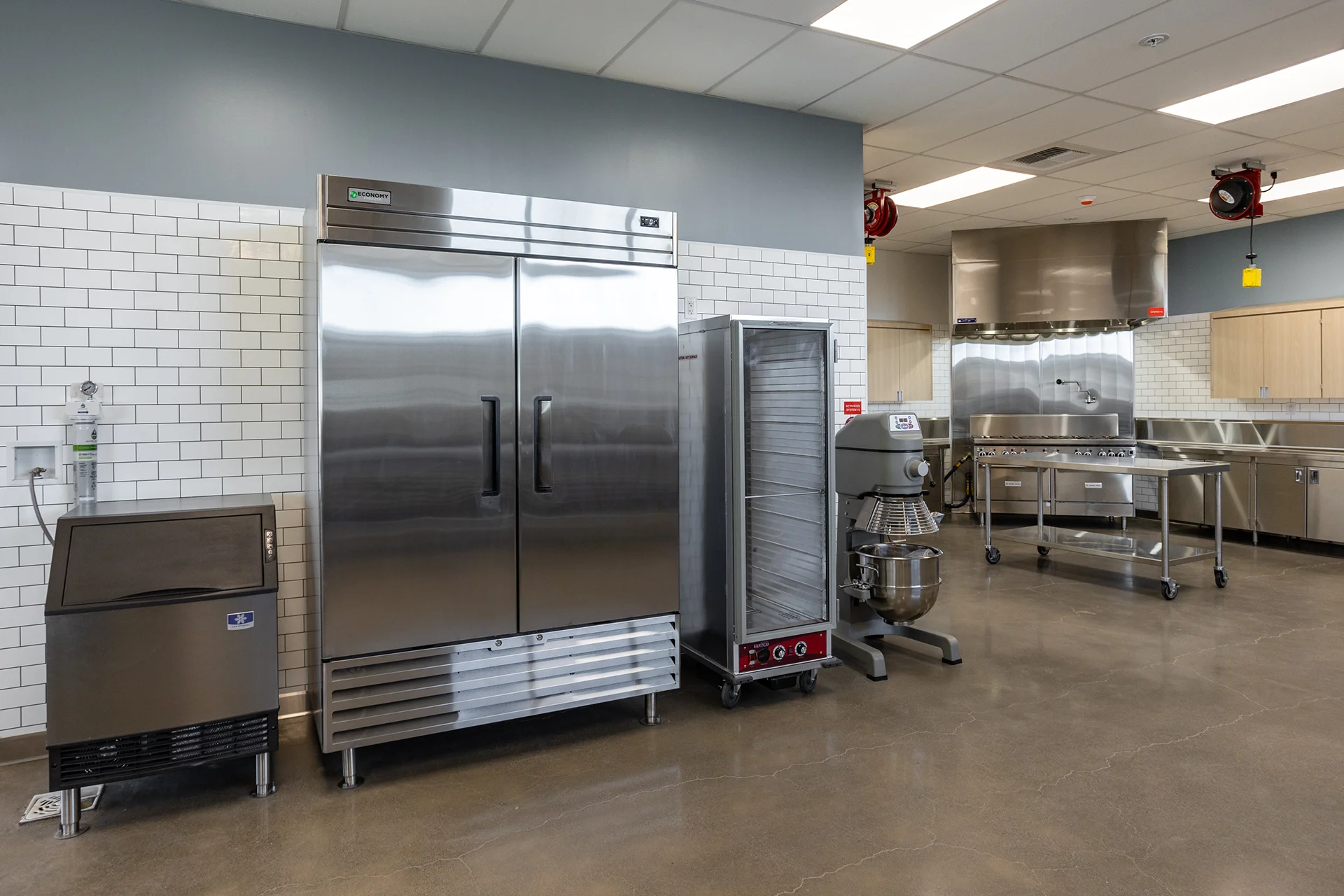 Commercial kitchen equipment alignment featuring a double-door stainless steel reach-in cooler, a mobile warming cabinet, a commercial floor mixer with a stainless steel bowl, and an industrial ice machine.
