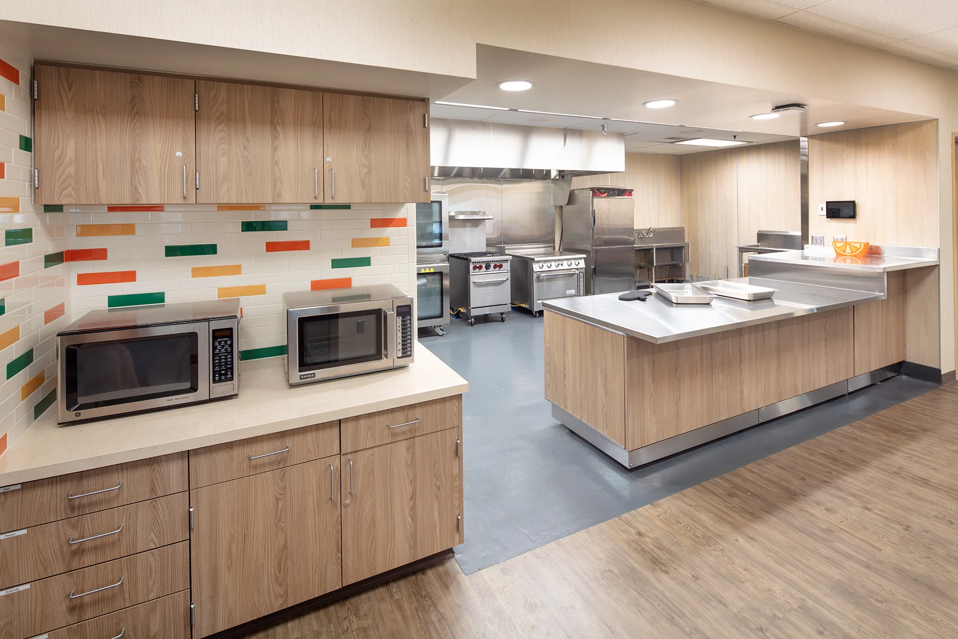 Commercial kitchen build out displaying stainless steel work tables, matching wood grain upper and lower cabinetry, commercial microwaves, a tiled subway tile backsplash, and professional vulcan cooking ranges over a seamless gray resin floor.