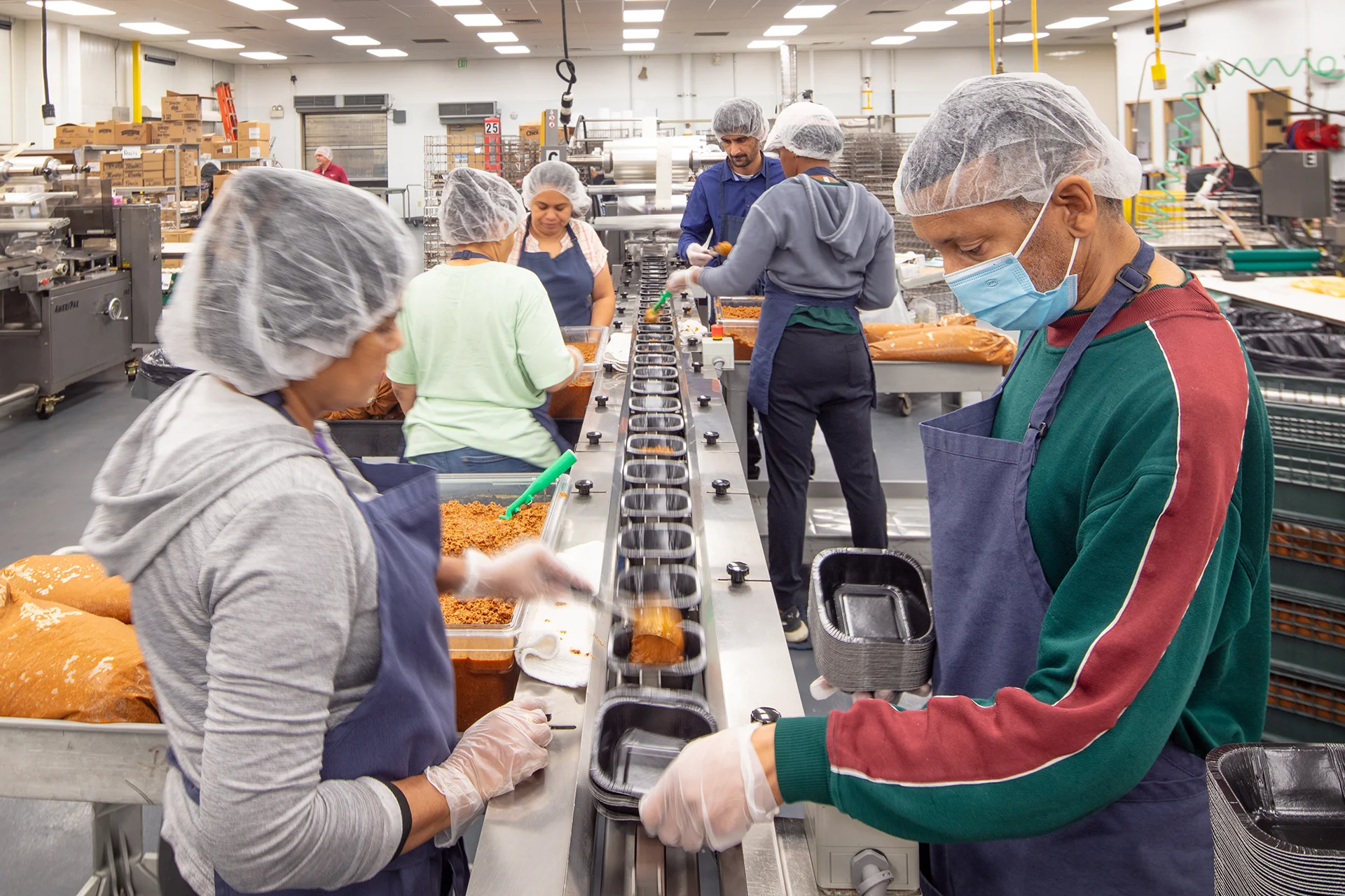 Commercial kitchen assembly station where staff in hairnets and face masks portion ingredients into containers along an automated food-grade stainless steel conveyor belt line.