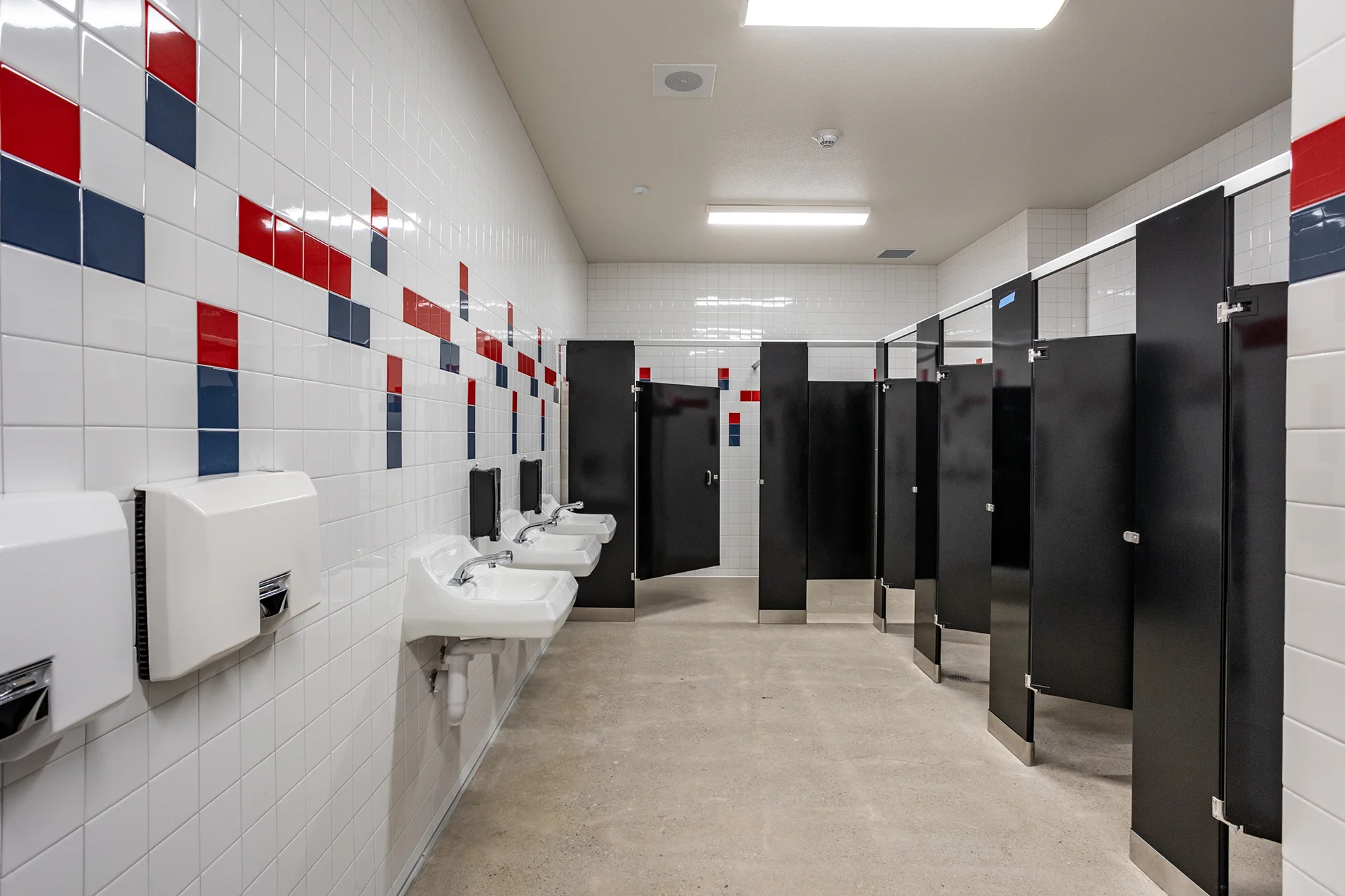 High school campus restroom renovation featuring clean white wall tiles accented by red and blue geometric patterns, multi-station wall-mounted porcelain sinks, heavy-duty commercial hand dryers, and black privacy stalls.