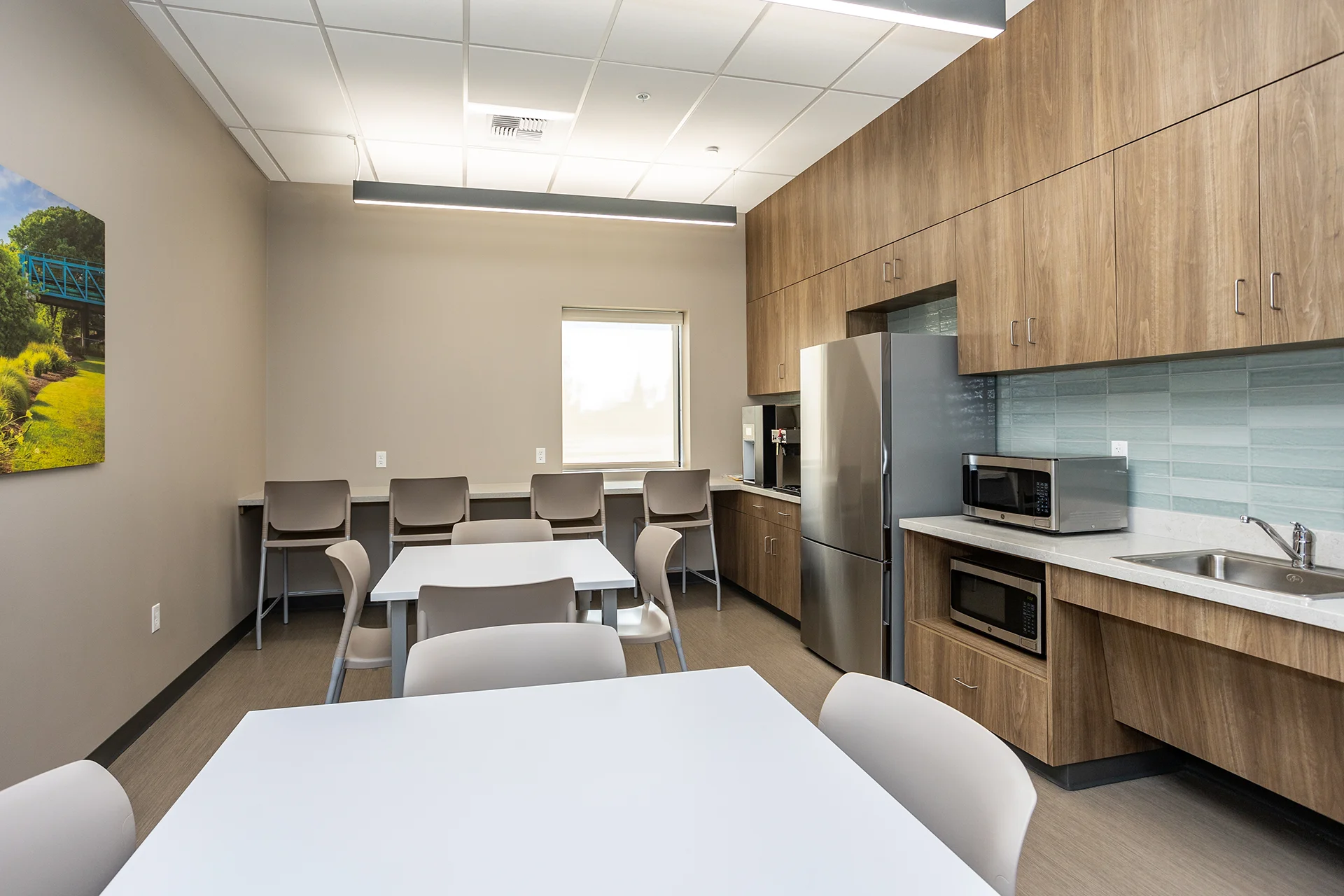 Modern employee dining room in Manteca, California, featuring white laminate lunch tables, grey molded plastic chairs, custom walnut-finish kitchen cabinetry, a stainless steel french-door refrigerator, a commercial glass tile backsplash, and built-in microwave alcoves.