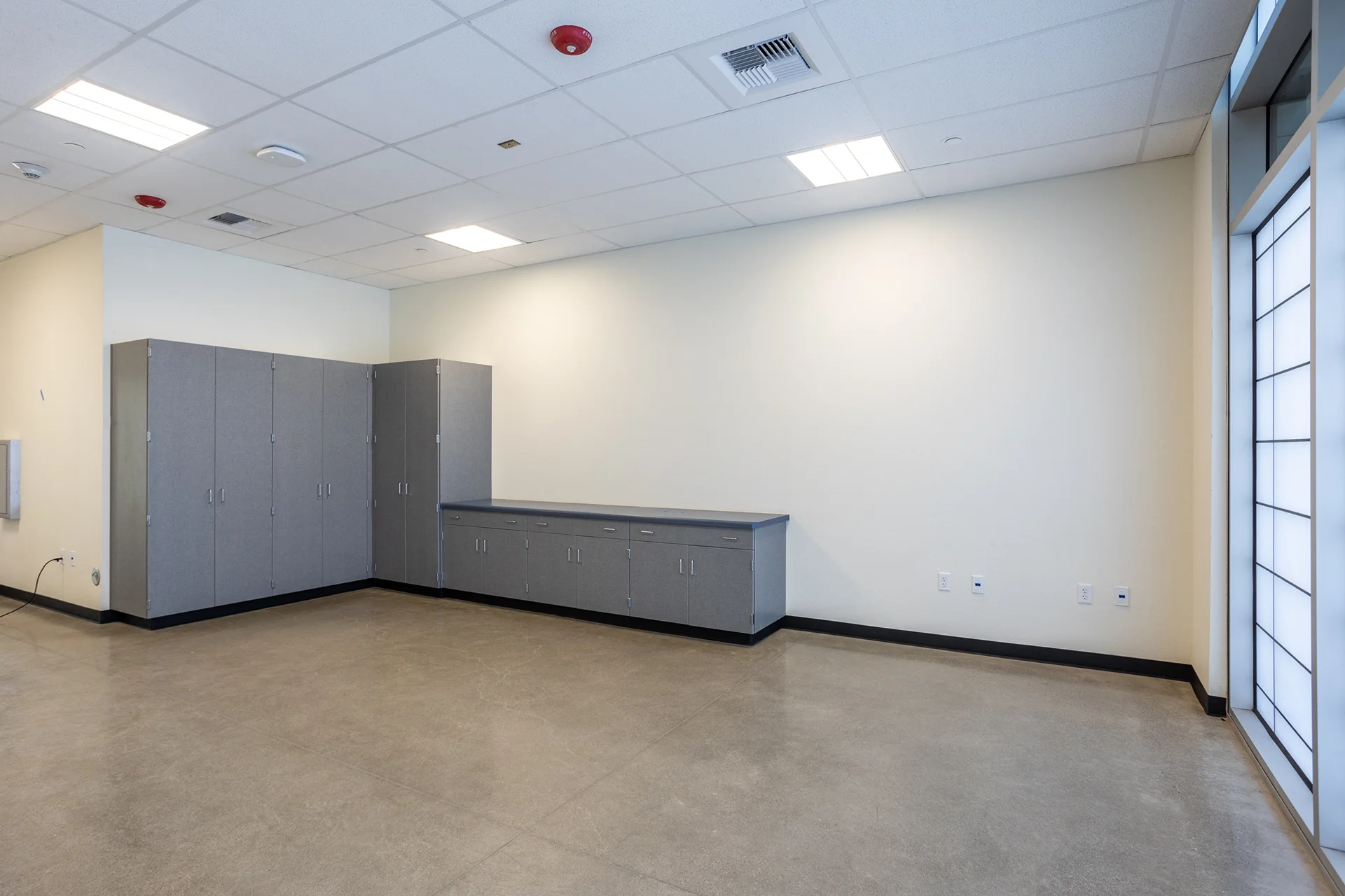 Academic campus storage facility showing heavy-duty gray laminate supply cabinets and base countertops, polished concrete floors, flat-panel LED lights, and a floor-to-ceiling frosted glass privacy window wall.
