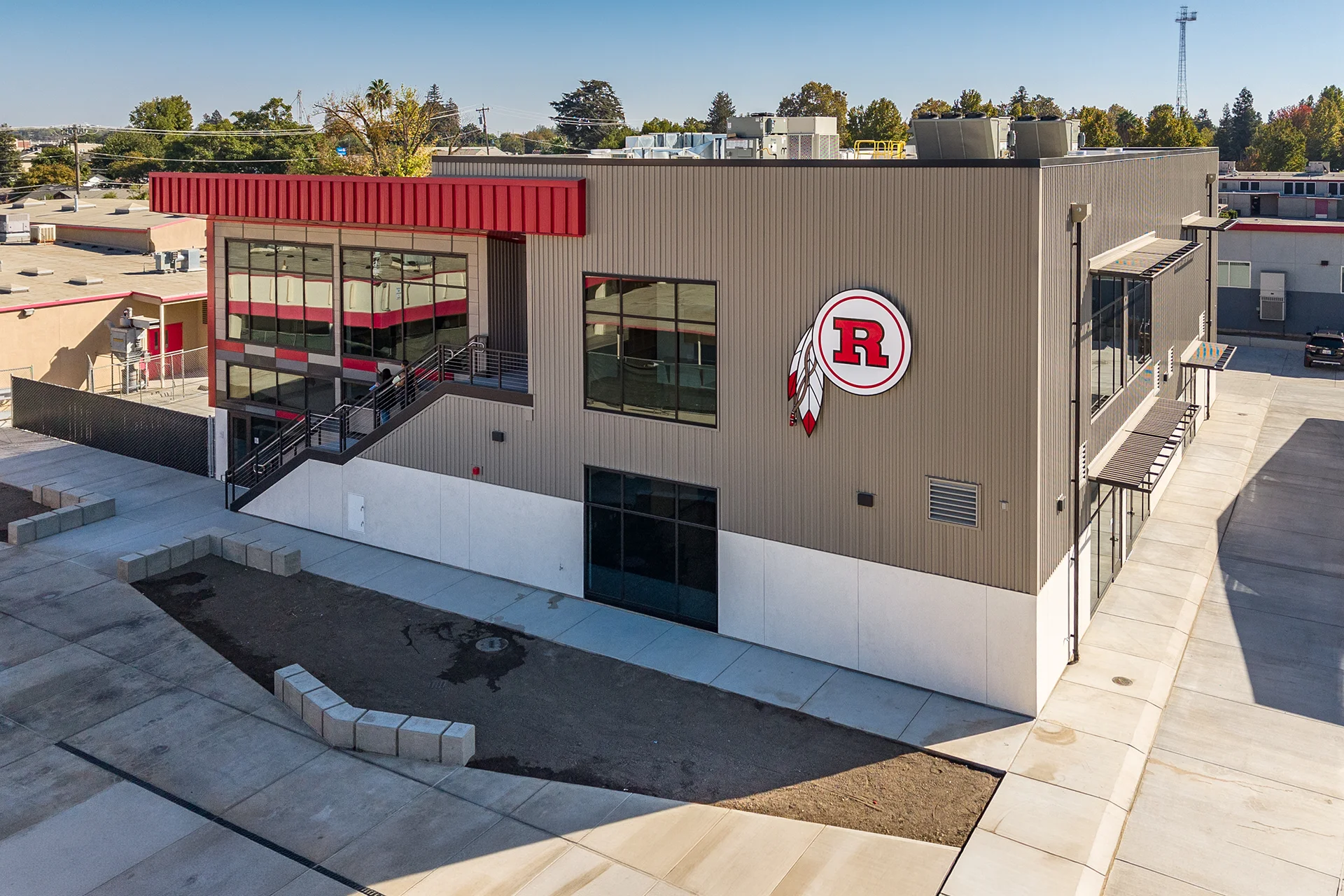 Elevated drone view of a contemporary two-story educational building engineered with gray vertical metal siding, an external concrete stairwell tower, a red metal accent canopy fascia, and angular geometric concrete site benches.