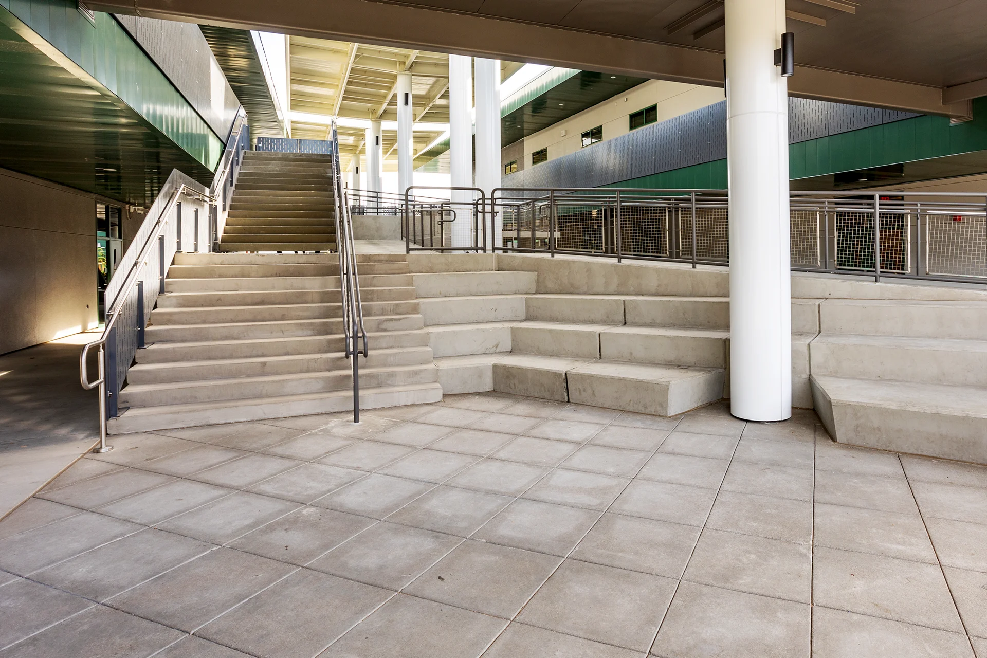 View of an outdoor educational gathering space in San Joaquin County, featuring wide cast concrete stairs with center steel handrails seamlessly transitioning into tiered concrete amphitheater seating beds beneath a large architectural metal canopy.