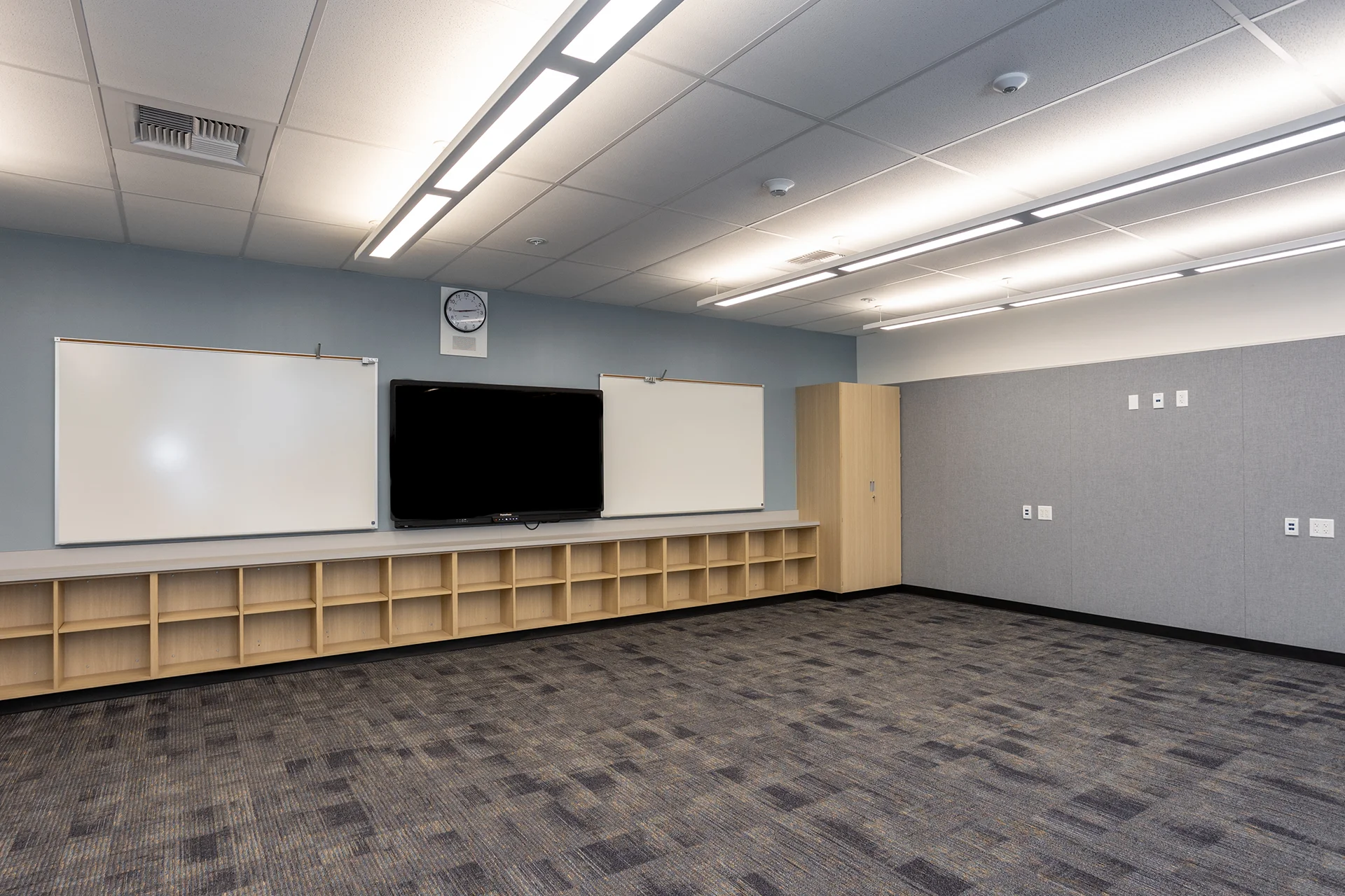 Angled view of a multi-purpose corporate or educational classroom highlighting a central interactive smart screen board, custom light-wood open shelving cubbies, flush-mounted acoustic wall panels, and gray geometric commercial carpet.