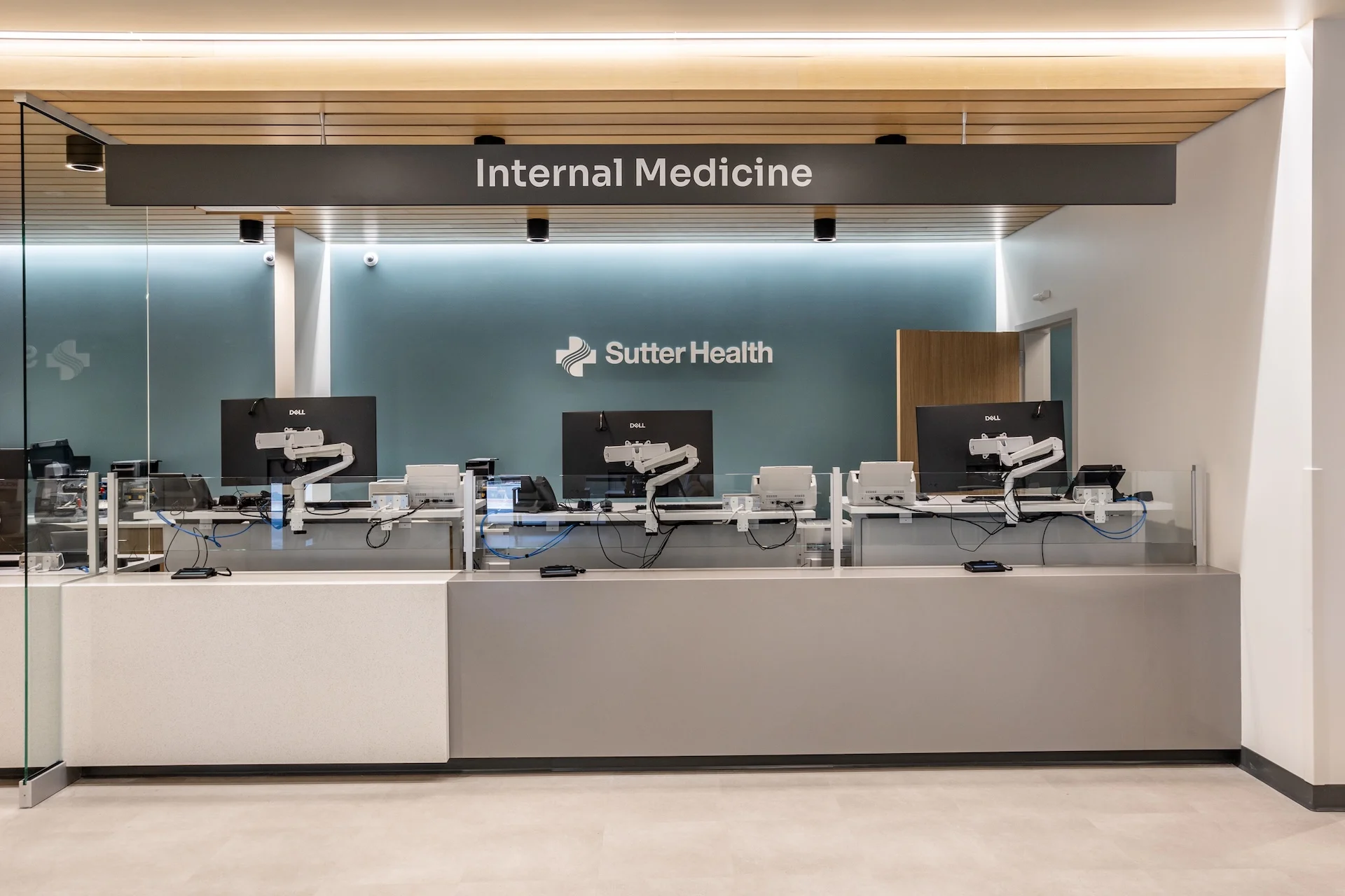 Clean architectural view of an internal medicine reception counter showcasing integrated wire-management raceways, ergonomic staff workstations, premium architectural glazing panels, and flush-mounted acoustic plaster ceilings with linear LED recess lighting.