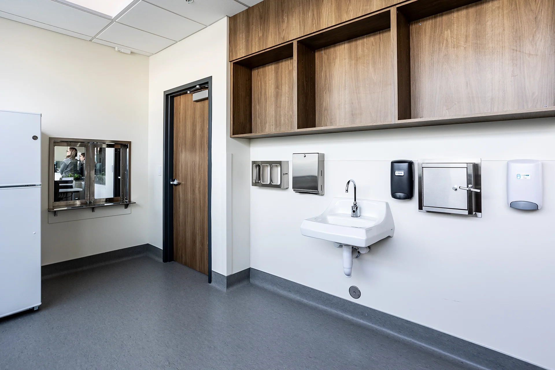 Detail view of a clinical specimen room showing a wall-hung ceramic handwashing sink with a goose-neck commercial faucet, an automated soap dispenser, a stainless steel paper towel holder, custom walnut wall cabinetry, and smooth grey commercial vinyl sheet flooring.