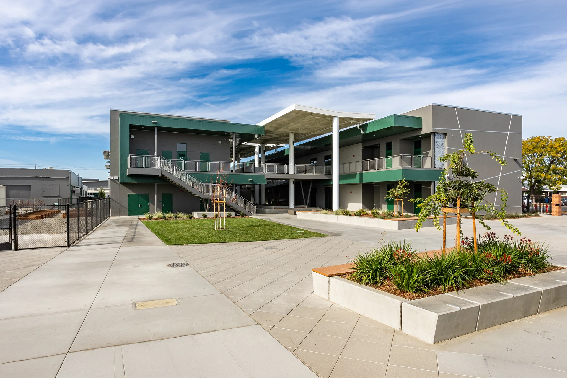 Expansive view of an educational plaza featuring modern two-story academic wings, a high-clearance architectural canopy, concrete walkways, a foreground concrete planter filled with ornamental grasses, and black steel security perimeter fencing.