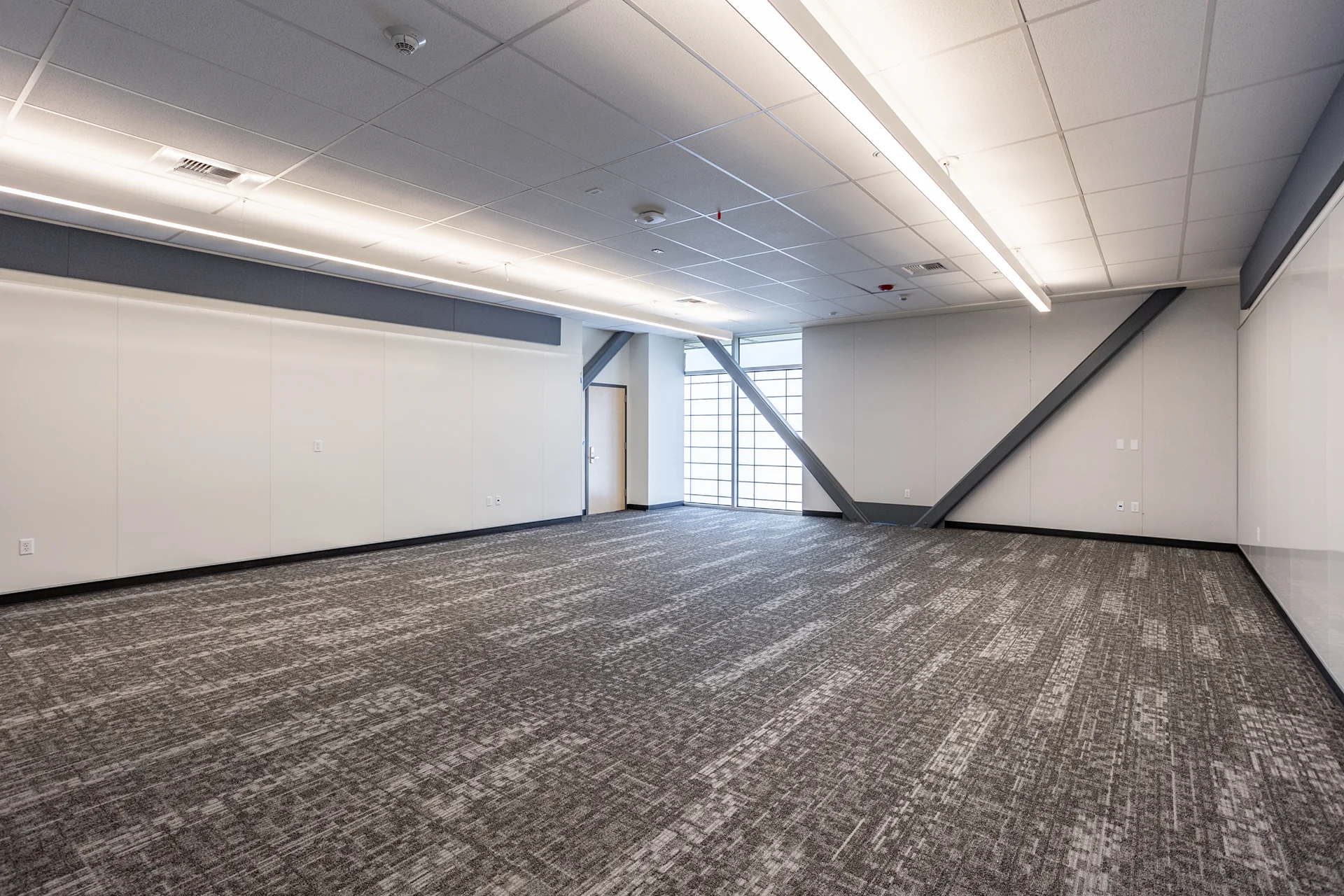 Expansive institutional learning hall showcasing industrial gray structural steel cross-bracing beams, high-performance patterned carpet tiles, sleek integrated whiteboards, and a floor-to-ceiling privacy window.