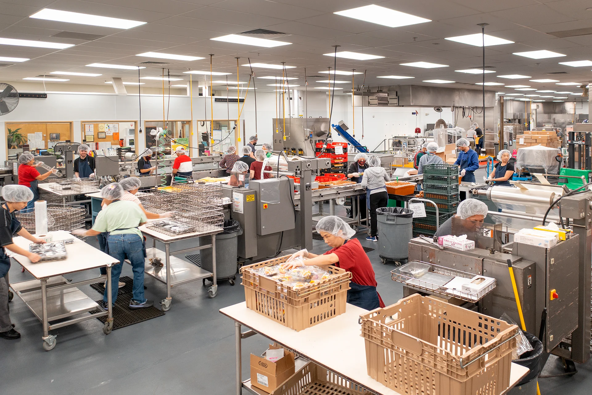 Wide-angle view of a multi-line institutional kitchen layout in Elk Grove, California, showcasing staff operating automated plastic wrapping machinery, custom stainless steel packing tables, and organized commercial distribution crates under high-efficiency drop-ceiling lighting.