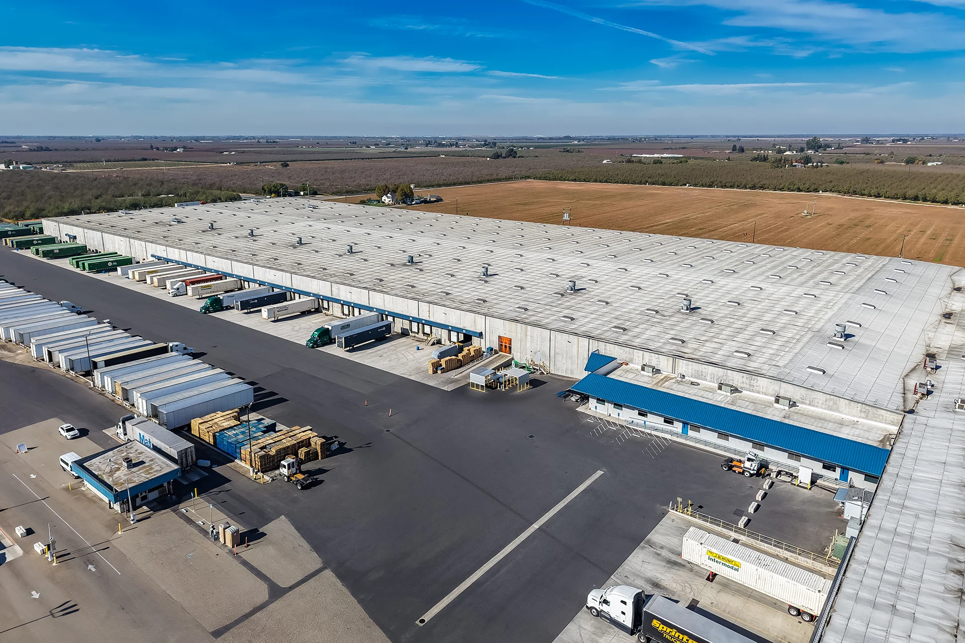 High-angle aerial drone view of a massive tilt-up concrete beverage distribution warehouse, showcasing a white low-slope single-ply roofing membrane with regular roof ventilators, recessed commercial loading docks with dock levelers, an asphalt staging yard filled with logistics trailers, and a secondary administrative office building with a blue metal canopy.