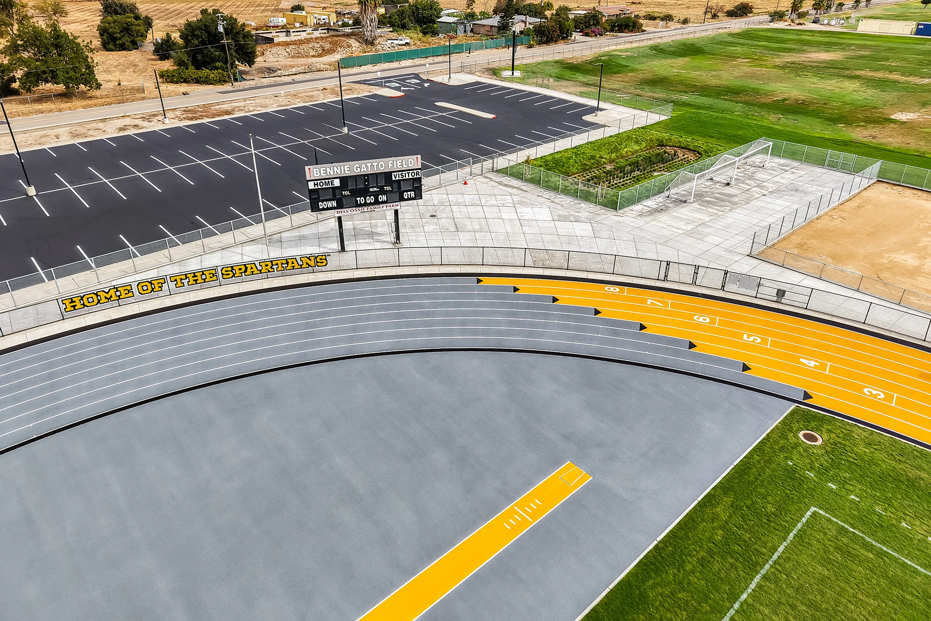 An aerial look at a synthetic running track curve next to the Bennie Gatto Field scoreboard and a newly paved asphalt parking lot.