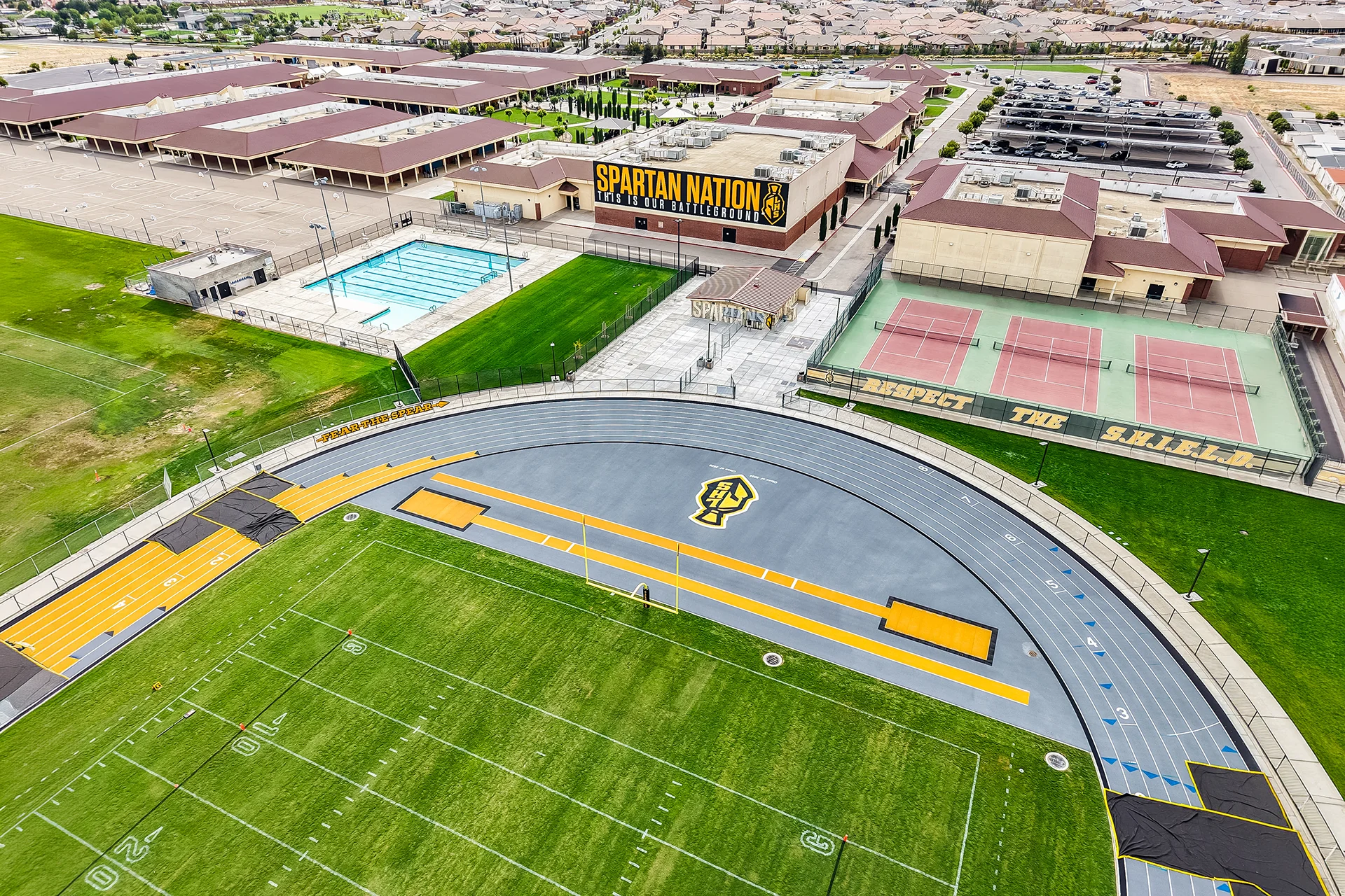 An expansive drone shot showcasing the Spartan Nation building mural, tennis courts, swimming pool, and the adjoining running track.